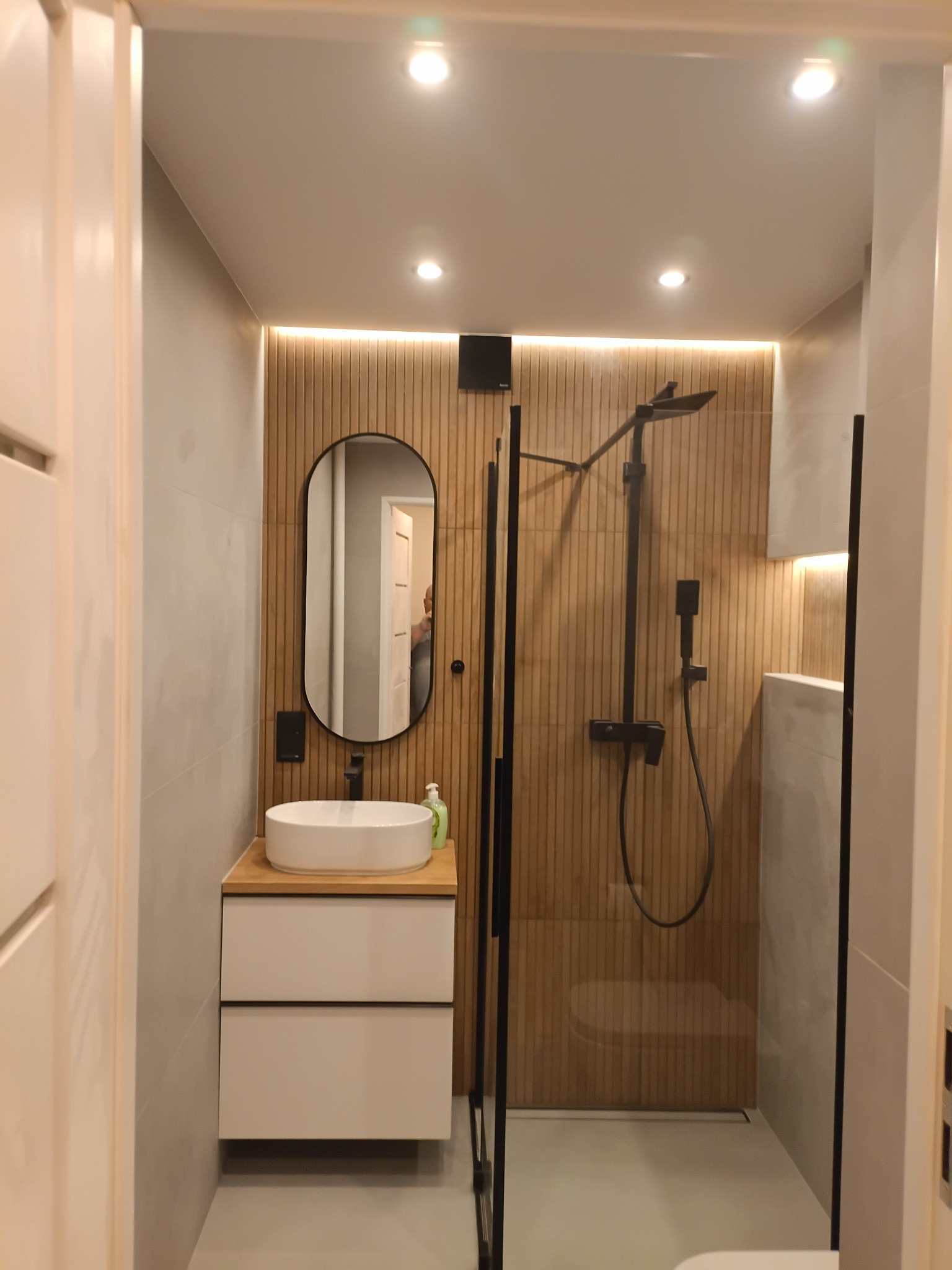 Nowoczesna łazienka z kabiną prysznicową typu walk-in, umywalką nablatową na szafce z szufladami i owalnym lustrem. Ściany wykończone płytkami i drewnianymi panelami.