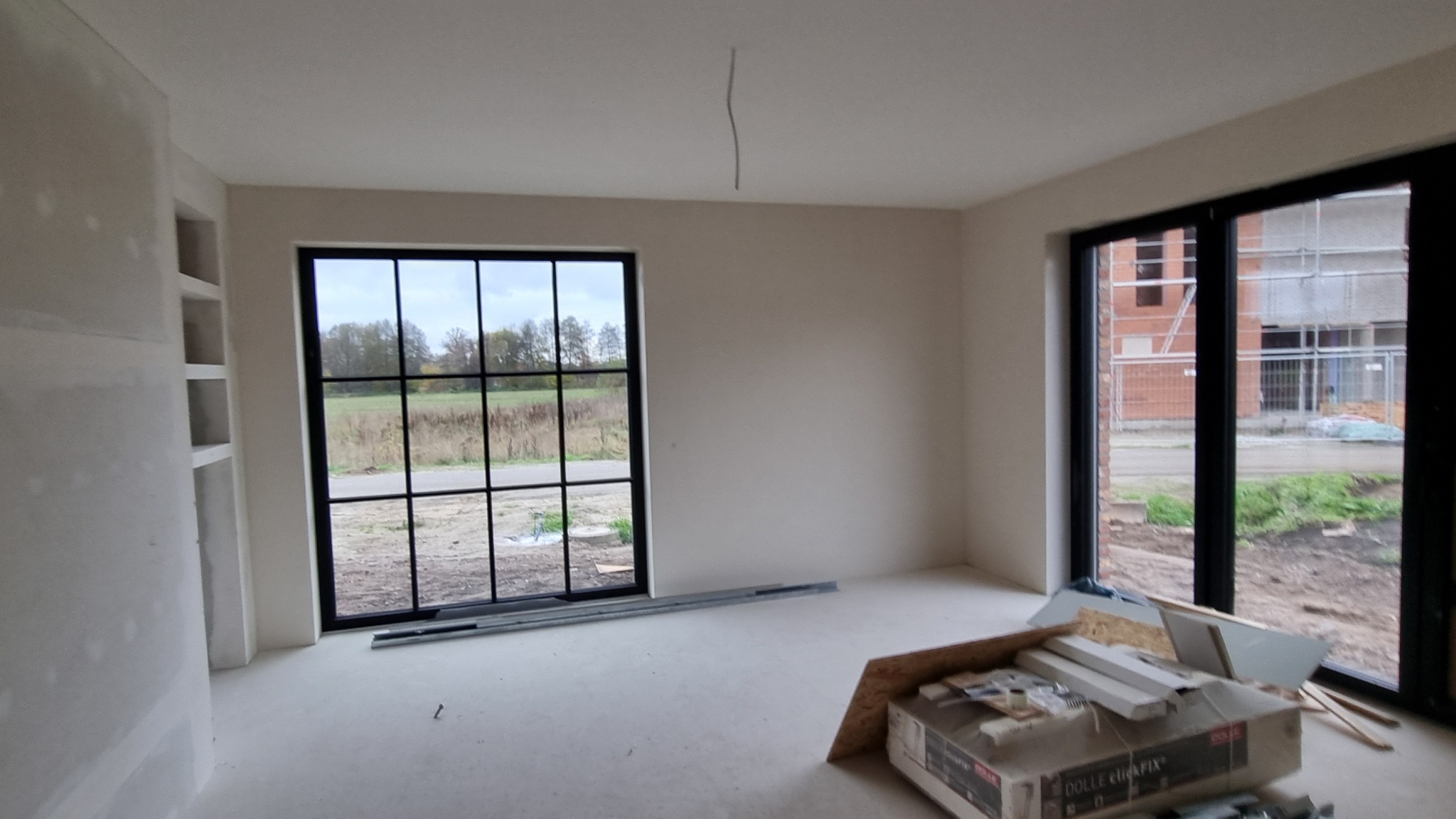 Surowe wnętrze w trakcie wykańczania, widoczne gładkie, białe ściany, duże okna z czarnymi ramami, materiały budowlane na podłodze i wnęka z półkami w ścianie.