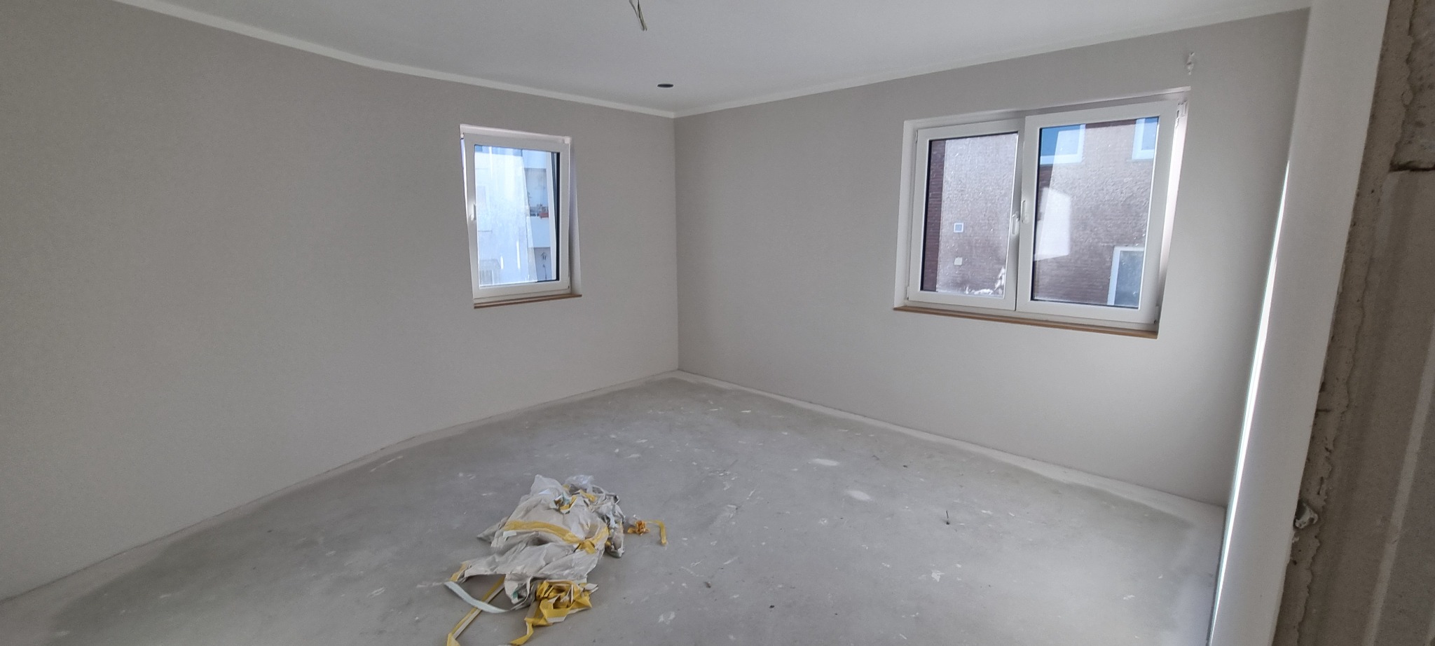 Pokój po wstępnym wykończeniu ścian i podłogi, widoczne dwie białe ramy okienne, szare ściany i podłoga w trakcie prac, na podłodze leżą resztki folii malarskiej.