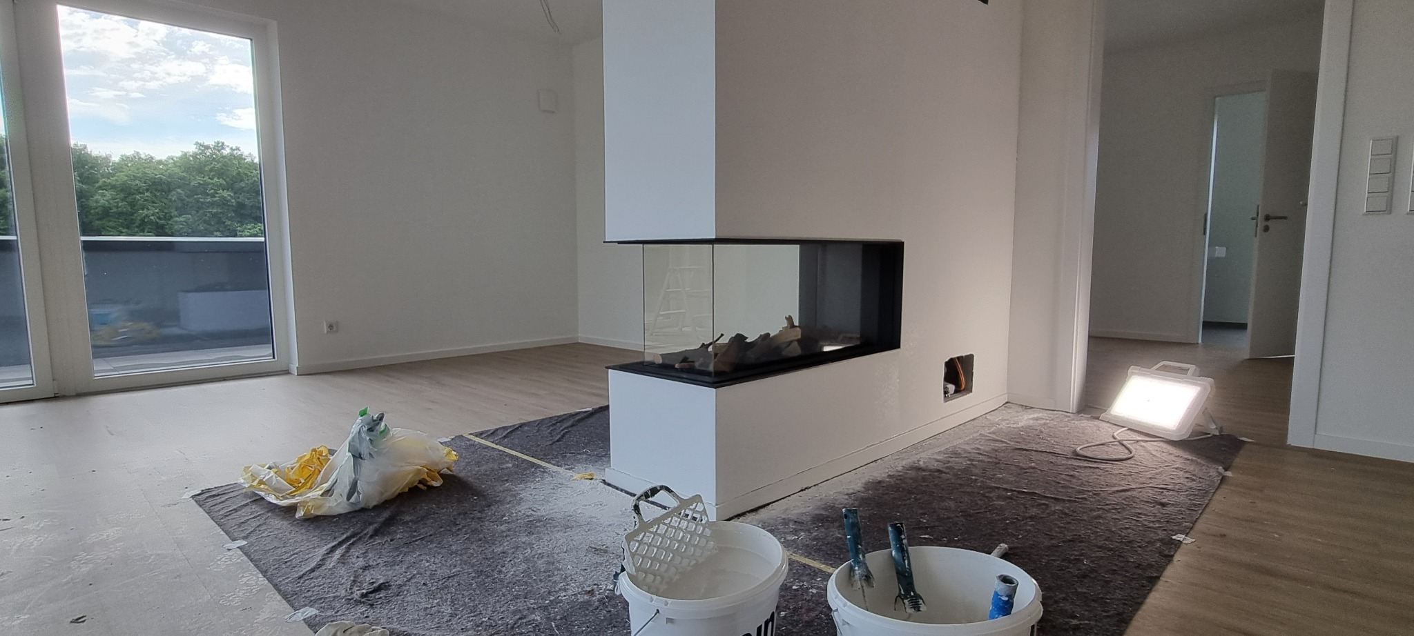 Wnętrze w trakcie prac wykończeniowych: zabezpieczona podłoga, wiaderka z farbą i wałki malarskie przy ścianie z nowoczesnym kominkiem; w tle duże okno balkonowe i otwarte drzwi do kolejnego...