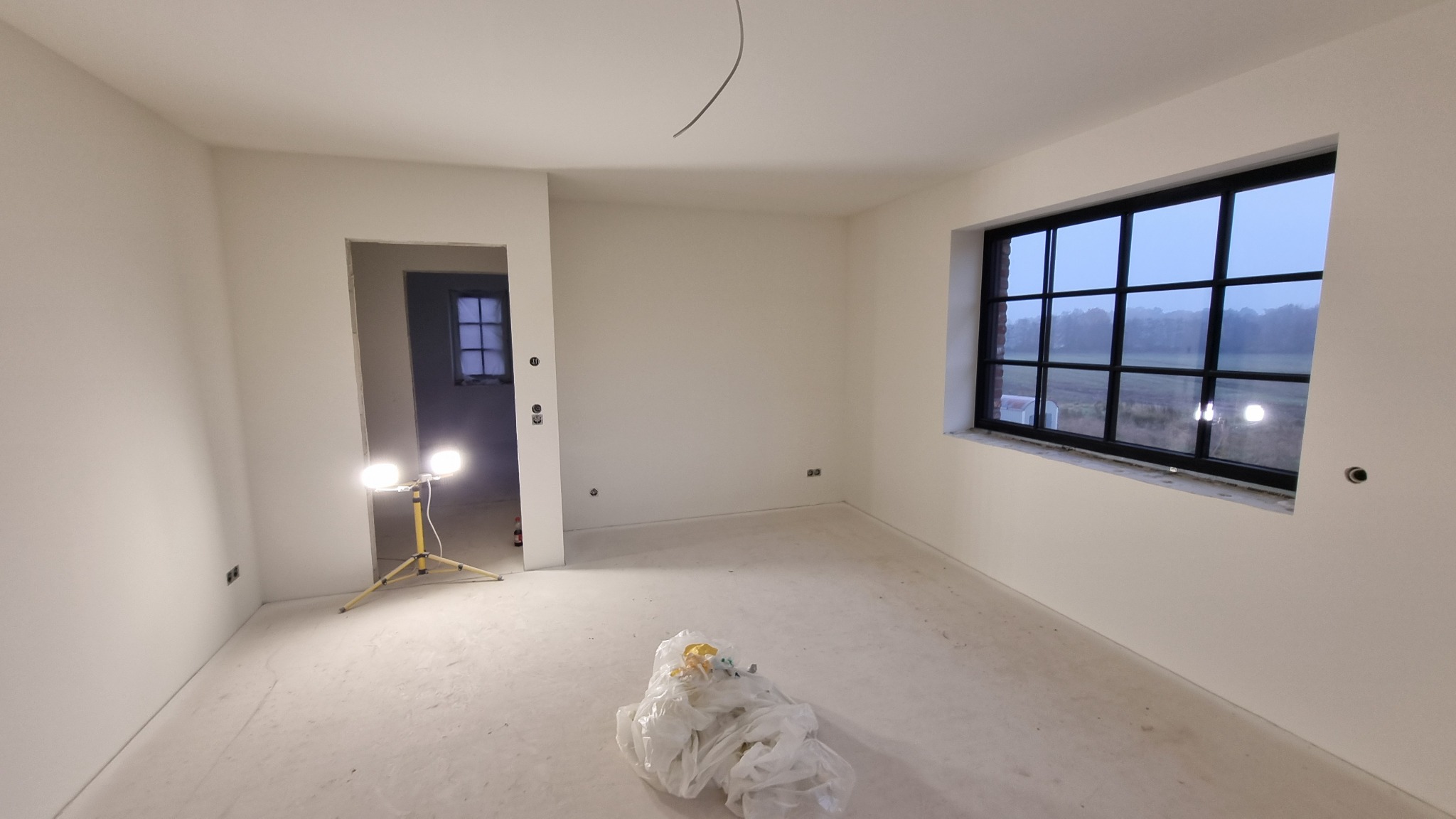 Pokój po szpachlowaniu i przygotowaniu do malowania, widoczna lampa halogenowa na statywie, worek z odpadami budowlanymi na podłodze i duże okno w czarnej ramie.