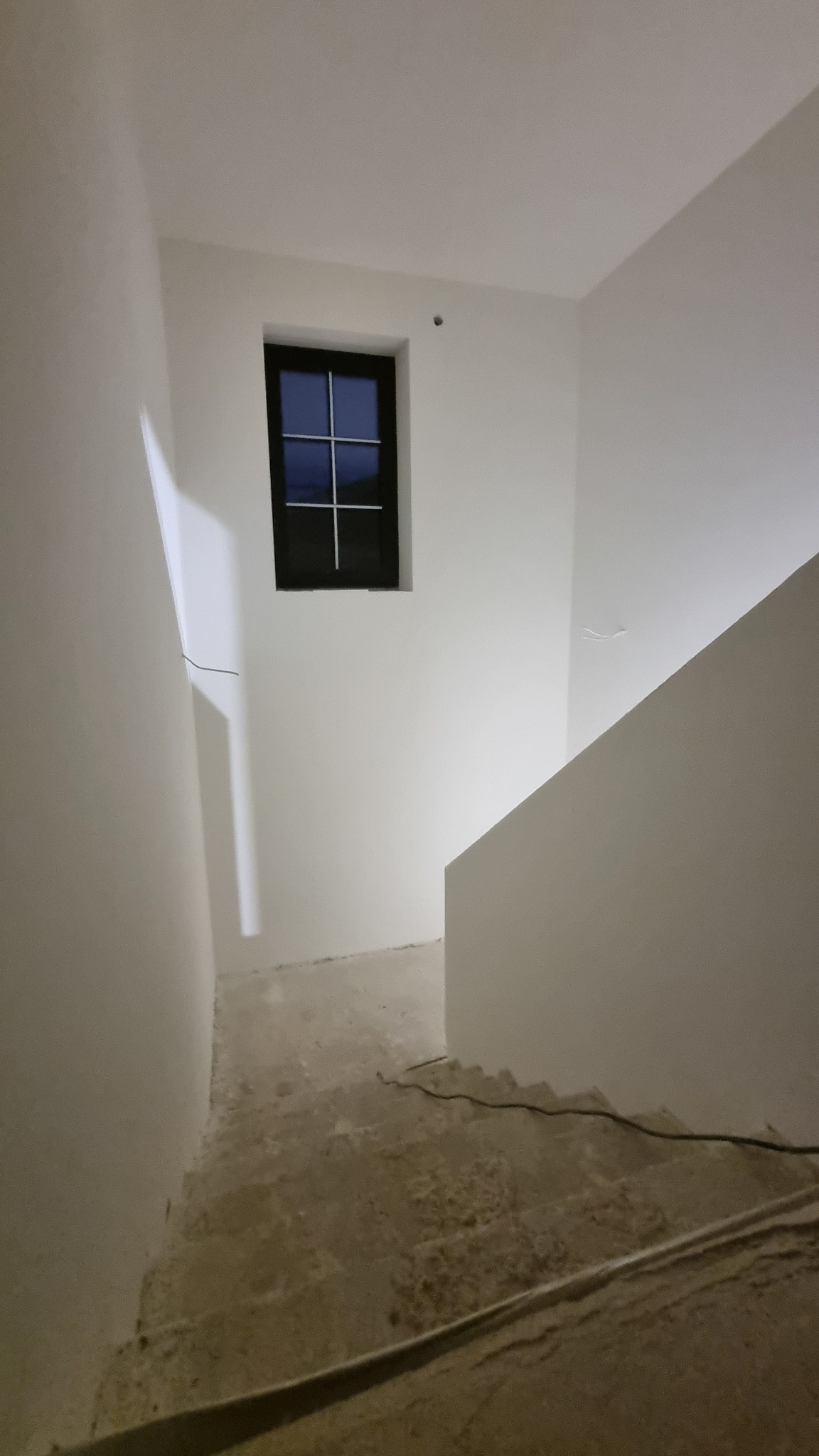 Wnętrze klatki schodowej w trakcie remontu, ściany świeżo pomalowane na biało, widoczne surowe schody i okno z czarną ramą.