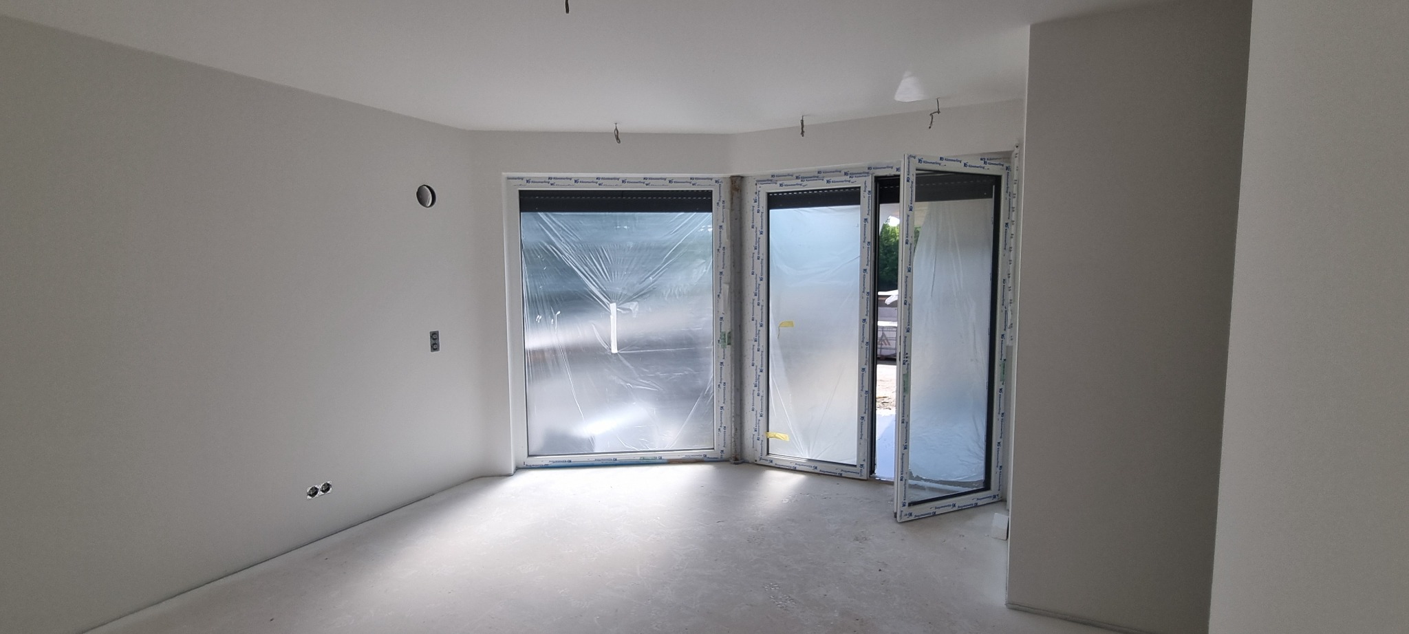 Wnętrze po szpachlowaniu i malowaniu na biało, okna zabezpieczone folią, widoczne gładkie ściany i surowa podłoga przed wykończeniem.