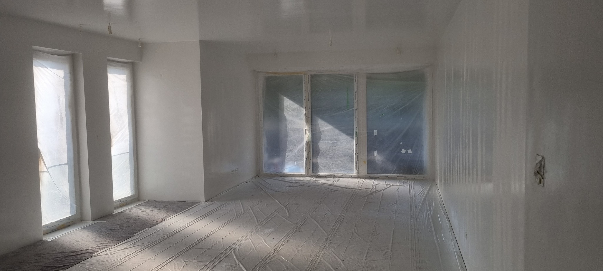 Pokój po świeżo wykonanych pracach, ściany i sufit pomalowane na biało, okna i podłoga zabezpieczone folią malarską, widoczne niedoskonałości na ścianach przed ostatecznym szlifowaniem.
