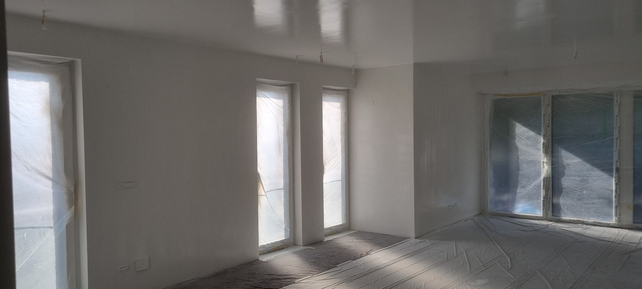 Wnętrze pokoju po nałożeniu gładzi, z widocznymi oknami zabezpieczonymi folią i połyskującą, gładką powierzchnią ścian i sufitu.
