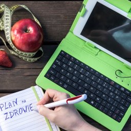 Julita Dudicz dietetyk dzieci i dorosłych - Poradnia PORCJA ZDROWIA - Plan na zdrowie zapisywany w notatniku, obok jabłko owinięte miarką krawiecką, truskawka, wizytówka i tablet z klawiaturą na drewnianym stole.