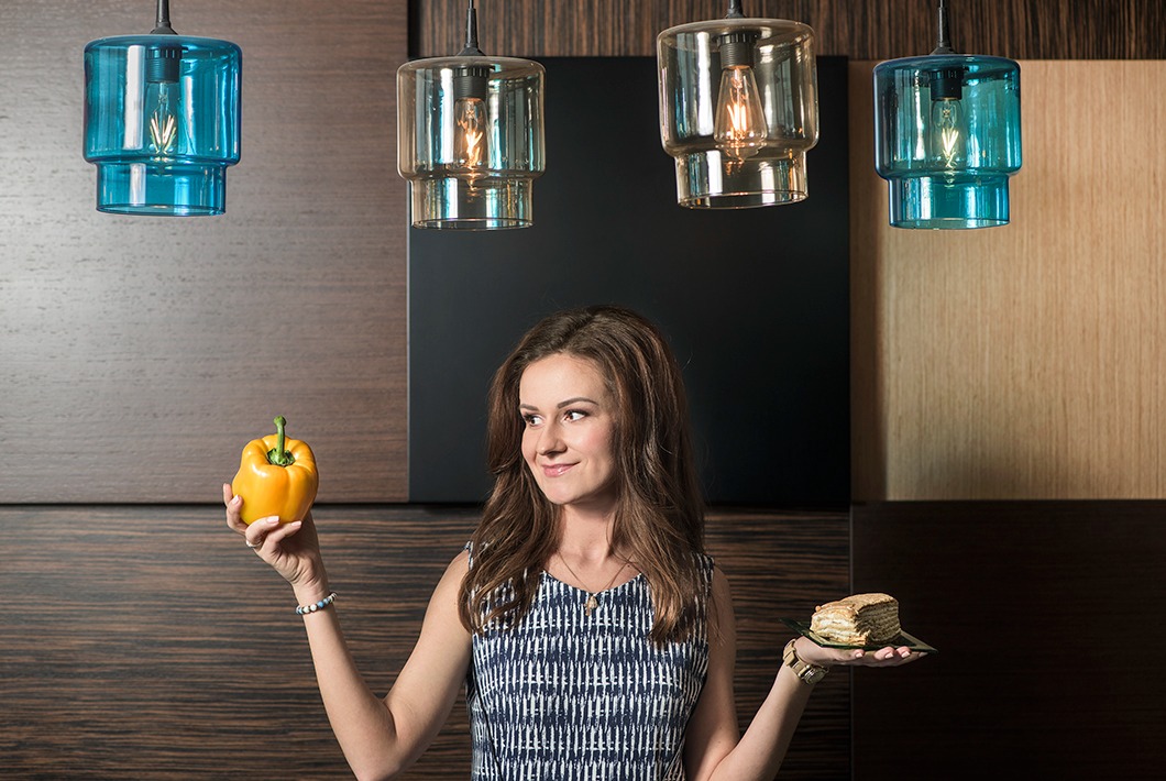 Uśmiechnięta kobieta trzymająca w jednej dłoni żółtą paprykę, a w drugiej kawałek tortu na małym talerzu, w tle wiszące lampy z żarówkami.