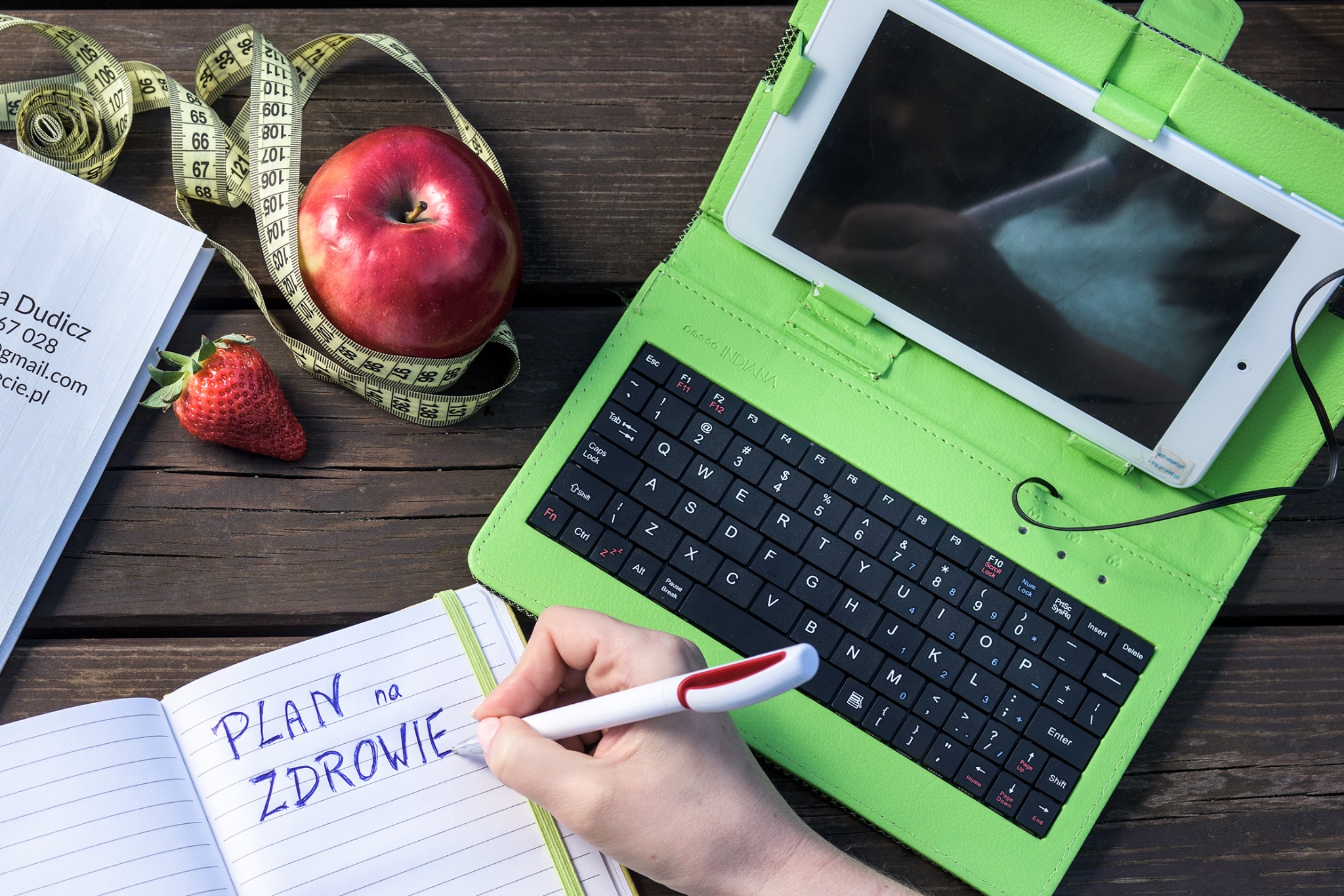 Plan na zdrowie zapisywany w notatniku, obok jabłko owinięte miarką krawiecką, truskawka, wizytówka i tablet z klawiaturą na drewnianym stole.