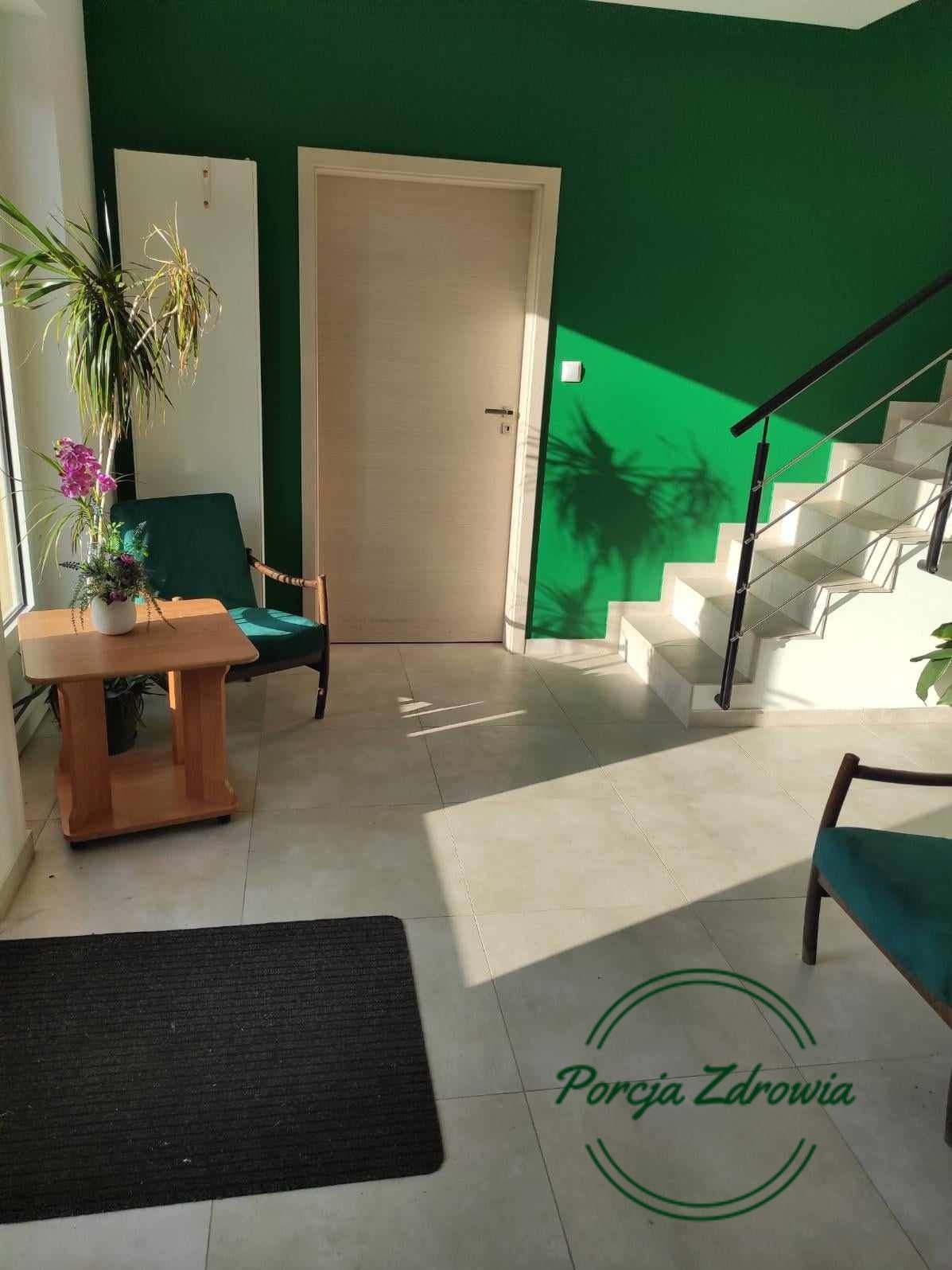 Jasny hol z zieloną ścianą, schodami i elementami wystroju: drewniany stolik z kwiatami, fotel w kolorze butelkowej zieleni, cień rośliny na ścianie. Na podłodze widoczne logo 'Porcja Zdrowia'.