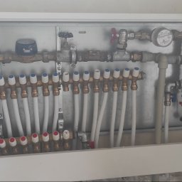 Usługi hydrauliczne Sokół - Rozdzielacz ogrzewania podłogowego w szafce, z widocznymi rurami i zaworami, zamontowany na ścianie z widocznymi rurami instalacyjnymi.