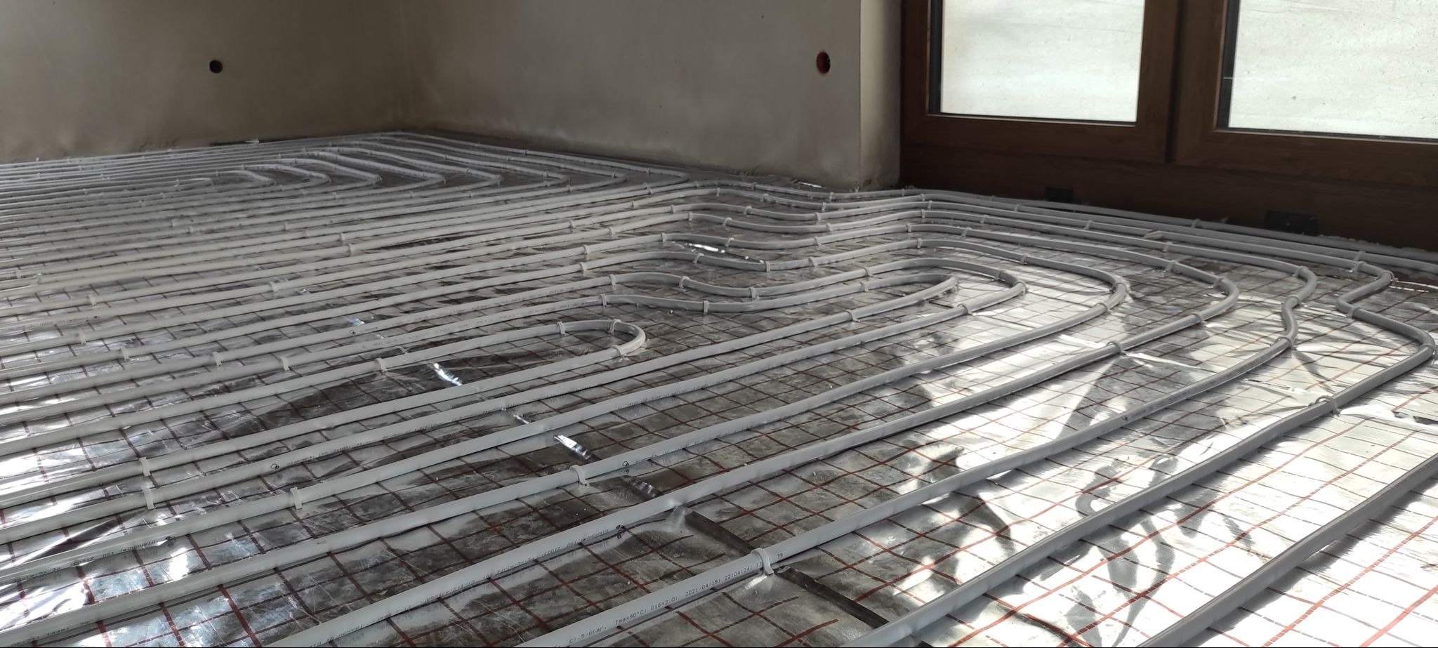 Szare rury ogrzewania podłogowego ułożone na srebrnej folii izolacyjnej w pomieszczeniu w Więcborku, widoczne mocowania i fragment okna.