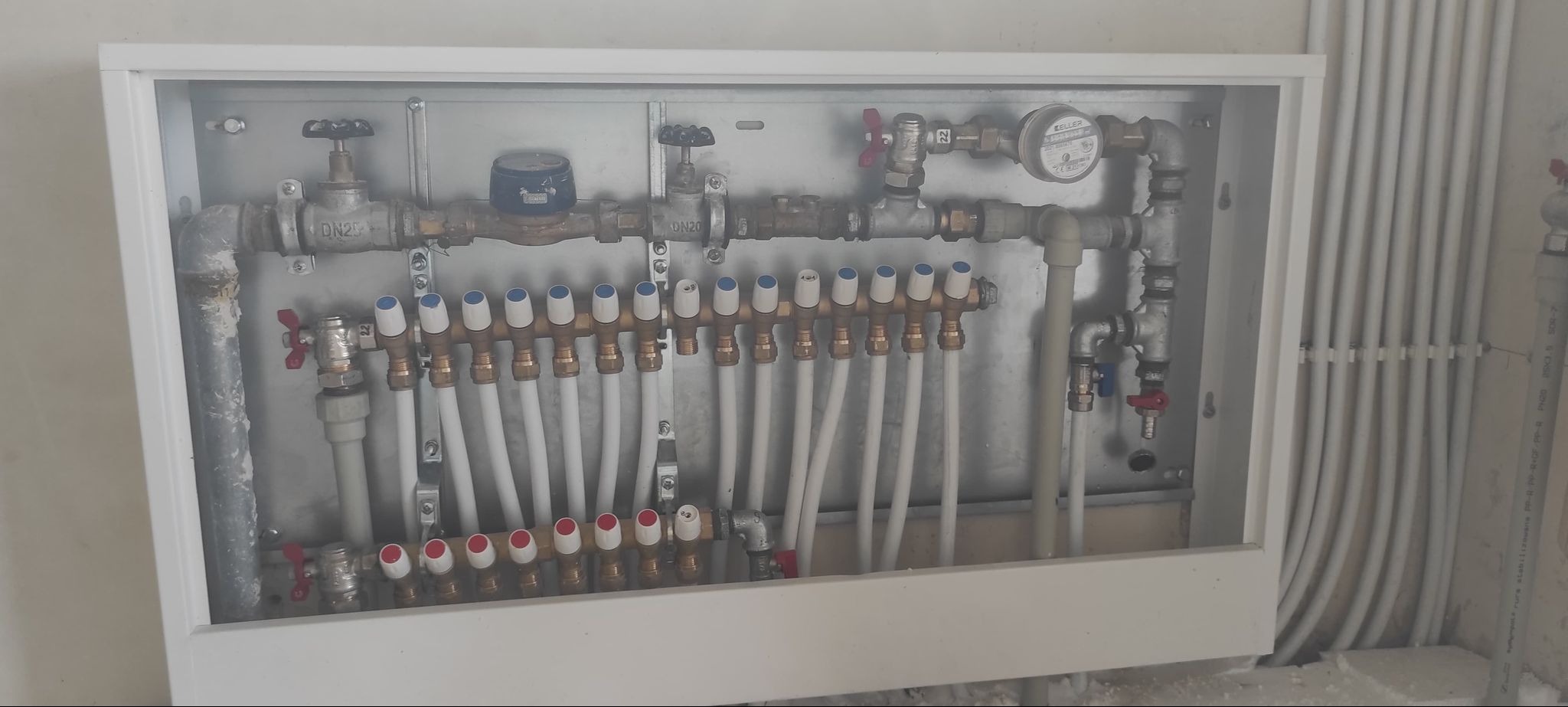 Rozdzielacz ogrzewania podłogowego w szafce, z widocznymi rurami i zaworami, zamontowany na ścianie z widocznymi rurami instalacyjnymi.