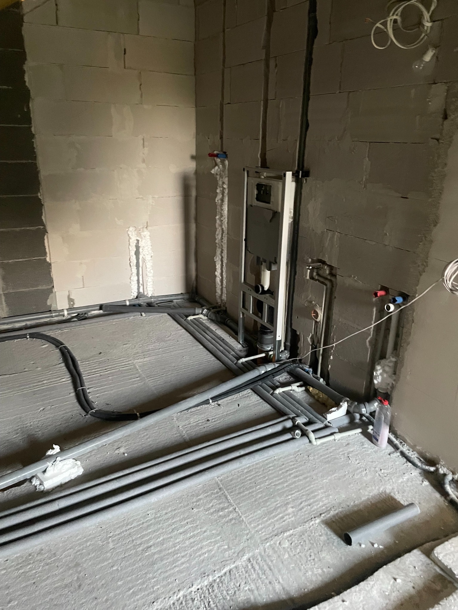 Surowe wnętrze w trakcie prac hydraulicznych: widoczne szare ściany z betonu komórkowego, liczne szare rury instalacyjne na podłodze i ścianach, stelaż podtynkowy WC zamontowany na ścianie...