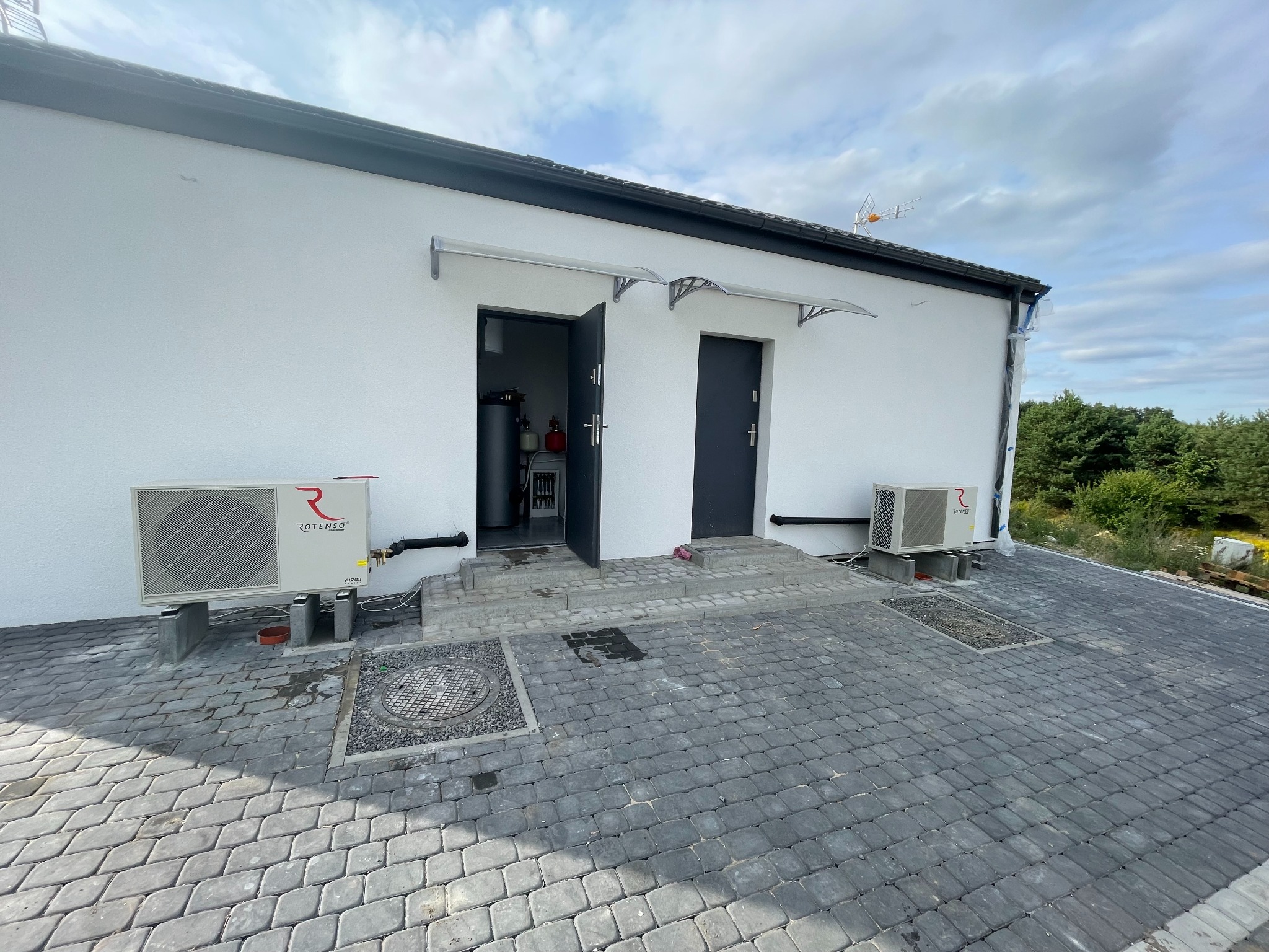 Biała ściana budynku z dwoma wejściami z zainstalowanymi klimatyzatorami marki Rotenso na zewnątrz, widoczne elementy instalacji CO w otwartym pomieszczeniu.