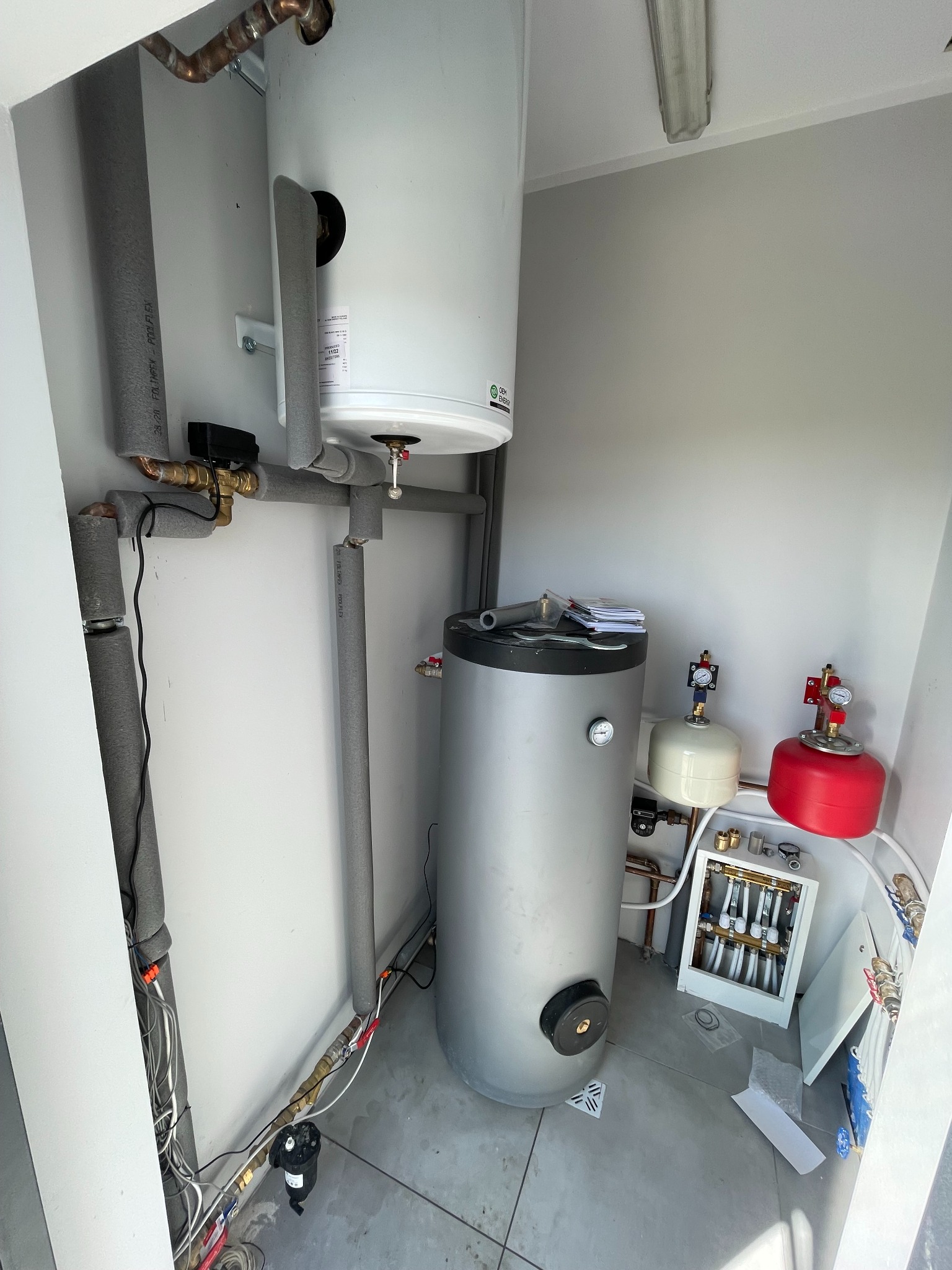 Dwa zbiorniki akumulacyjne ciepłej wody, biały i srebrny, połączone rurami w kotłowni. Widoczny rozdzielacz ogrzewania podłogowego, zawory, manometry i izolowane rury. Część instalacji w trakcie...