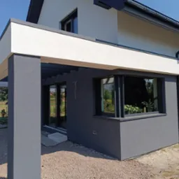 Nowoczesny dom z szarą elewacją i białym wykończeniem, widoczne okno z roślinami wewnątrz, żwirowe podłoże.