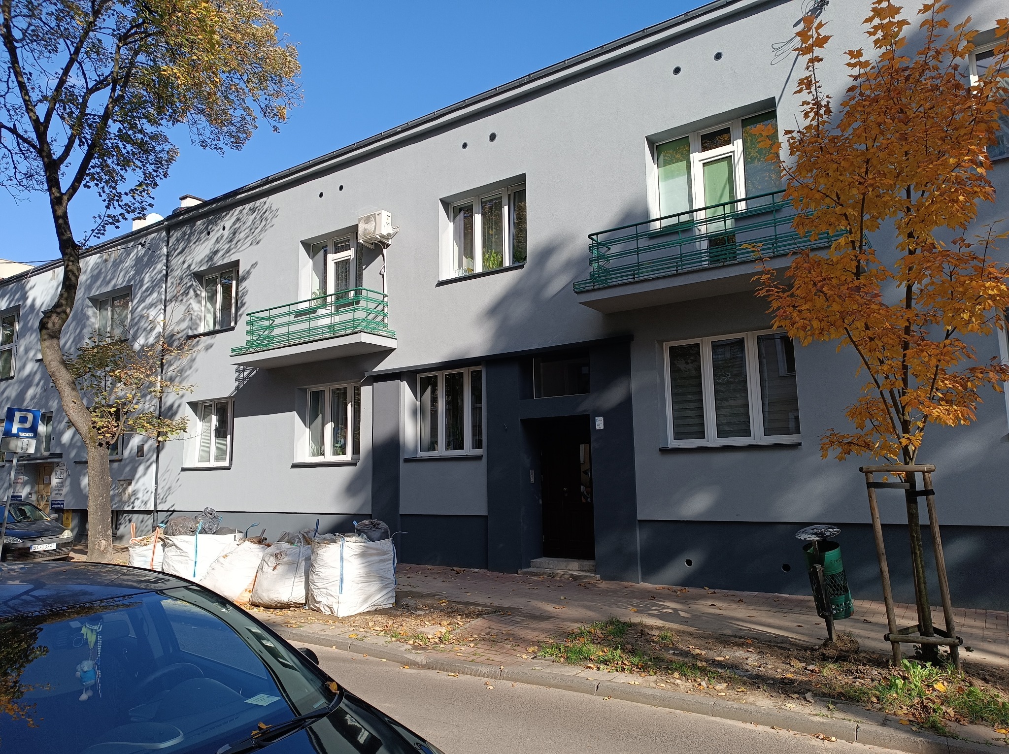 Szary budynek wielorodzinny z balkonami w stylu art deco, widoczne worki z odpadami budowlanymi na chodniku, drzewo z żółtymi liśćmi i fragment czarnego samochodu.