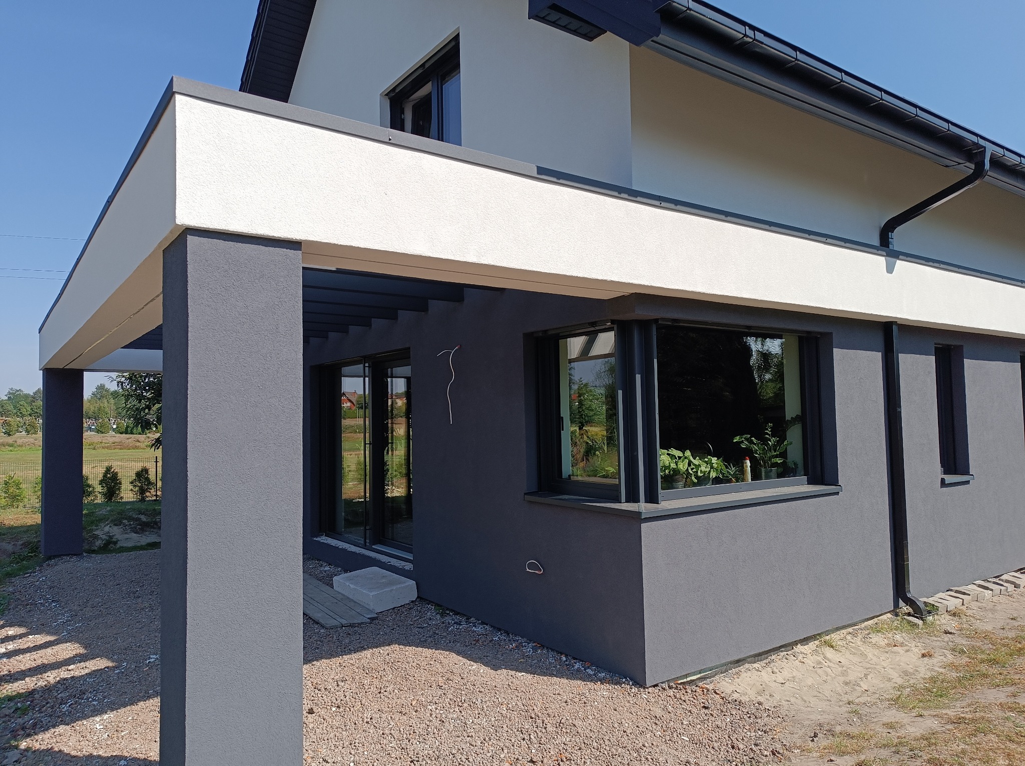 Nowoczesny dom z szarą elewacją i białym wykończeniem, widoczne okno z roślinami wewnątrz, żwirowe podłoże.