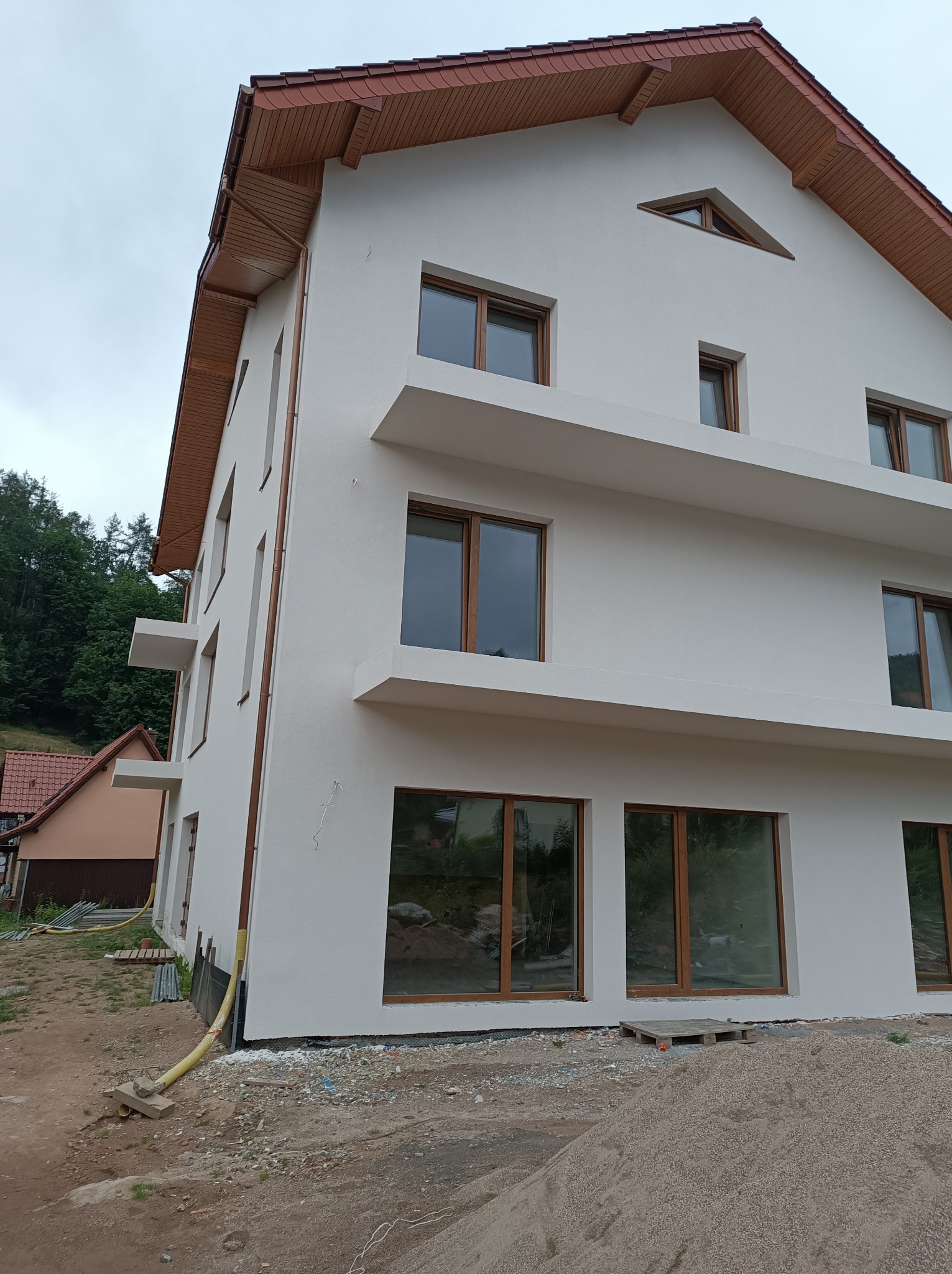 Nowoczesny, trzypiętrowy budynek z białą elewacją i drewnianymi oknami, z widocznymi elementami konstrukcyjnymi w formie poziomych półek, na tle zieleni i sąsiedniego domu w trakcie prac...