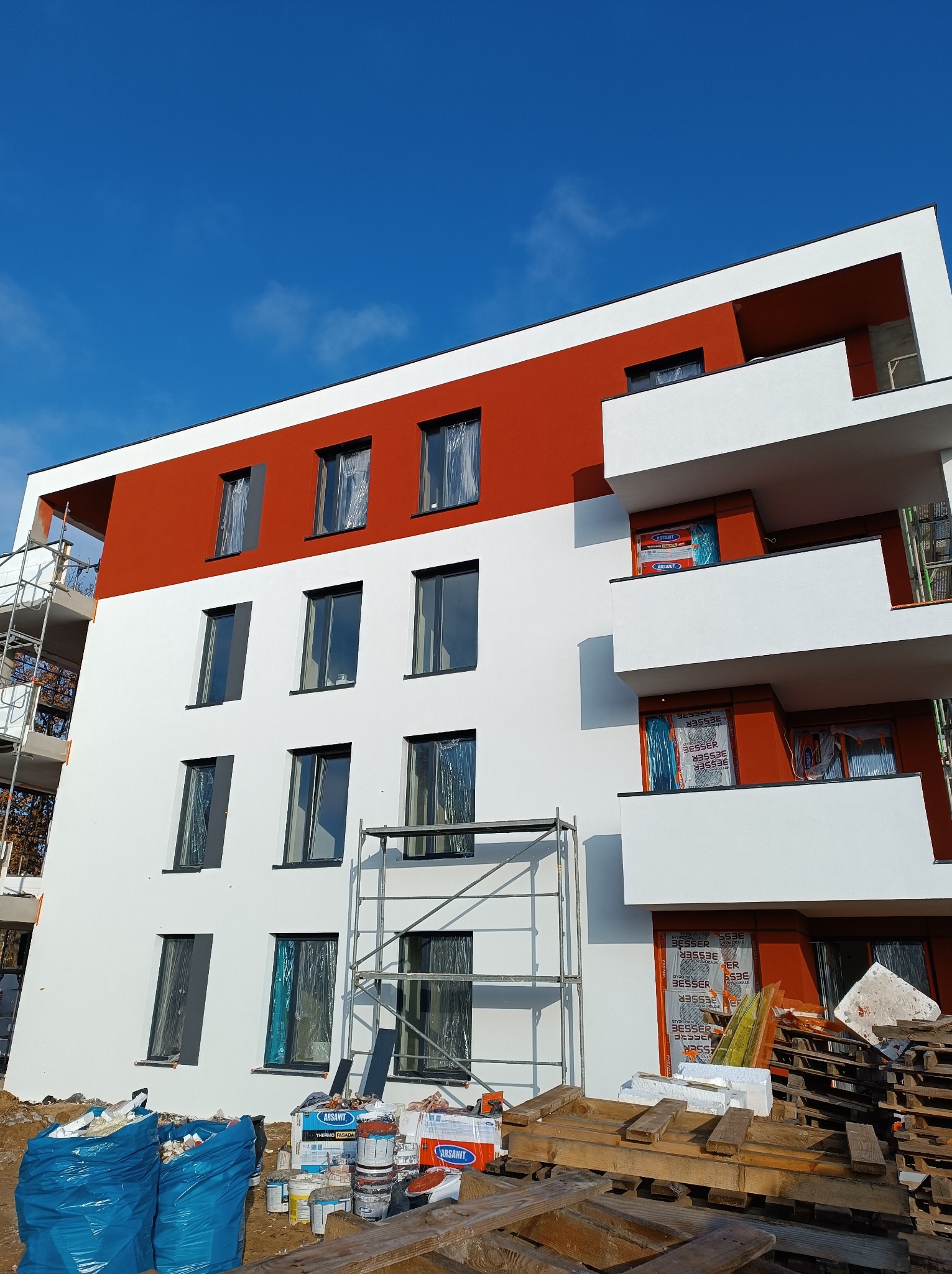 Nowoczesny budynek w Żarkach z częściowo wykonaną elewacją w kolorze białym i czerwonym, widoczne rusztowania, materiały budowlane i zabezpieczone okna folią.