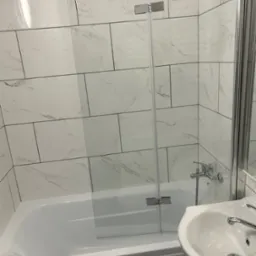 Nowoczesna łazienka z białą armaturą i szklanym parawanem nawannowym, płytki imitujące marmur na ścianach, widoczny fragment umywalki i baterii.