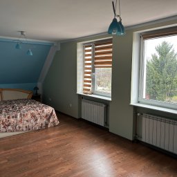 Kacper Markuszewski - Pokój na poddaszu z drewnianą podłogą, łóżkiem z wzorzystą narzutą, oknami z widokiem na zieleń i grzejnikami pod oknami. Ściany w kolorze zielonym i niebieskim.