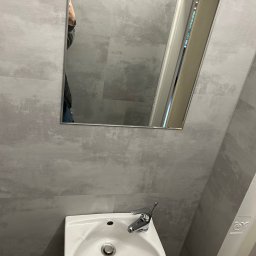 Kacper Markuszewski - Mała łazienka z umywalką i lustrem, szare płytki imitujące beton na ścianach, minimalistyczna armatura.