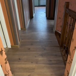 Kacper Markuszewski - Długi korytarz z panelami podłogowymi w odcieniach brązu, ściany w paski i z kwiatowym wzorem, widok na okno na końcu korytarza.