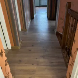 Długi korytarz z panelami podłogowymi w odcieniach brązu, ściany w paski i z kwiatowym wzorem, widok na okno na końcu korytarza.