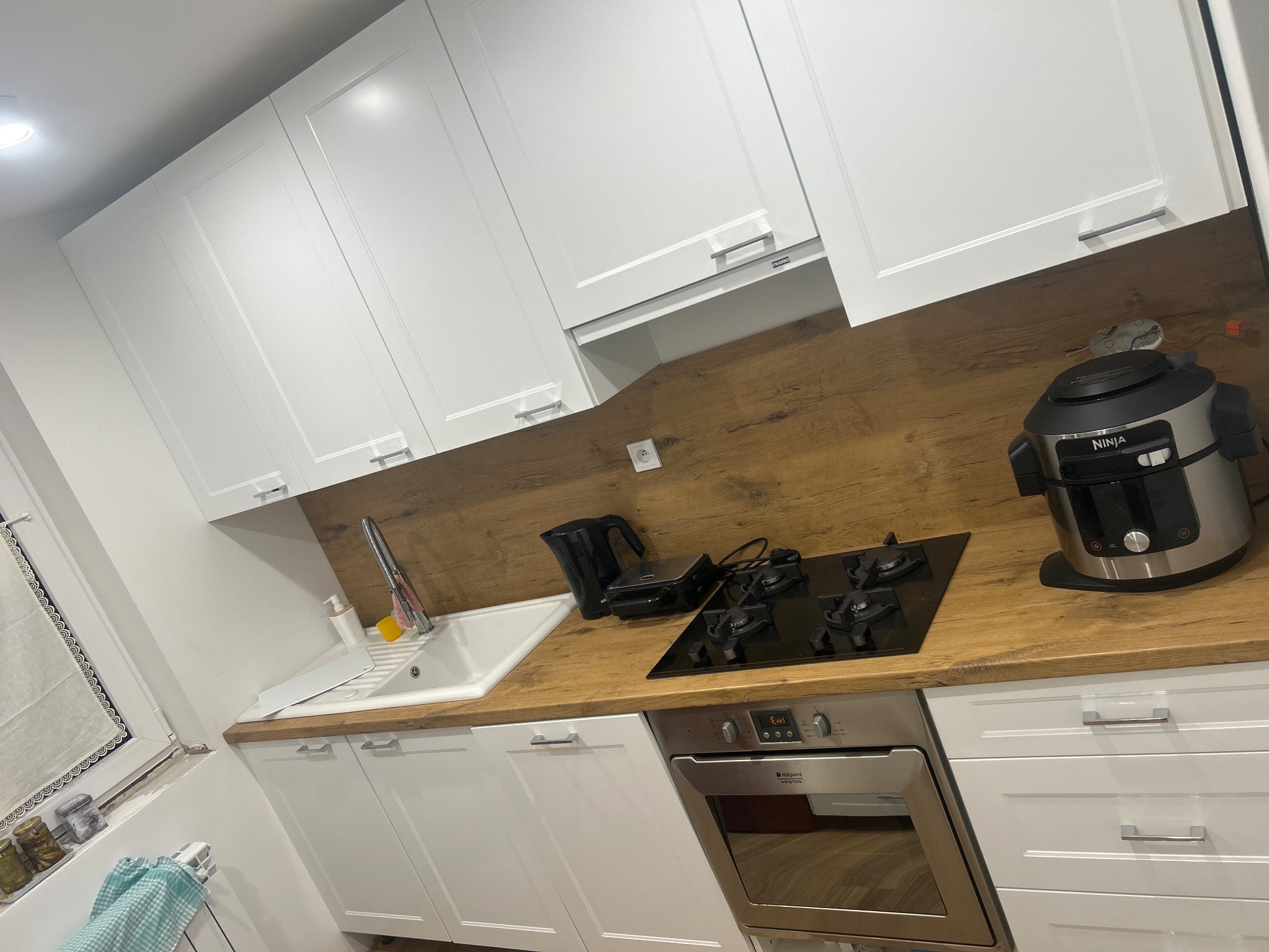 Nowoczesna kuchnia z białymi szafkami, drewnianym blatem i czarną płytą grzewczą oraz piekarnikiem Hotpoint Ariston