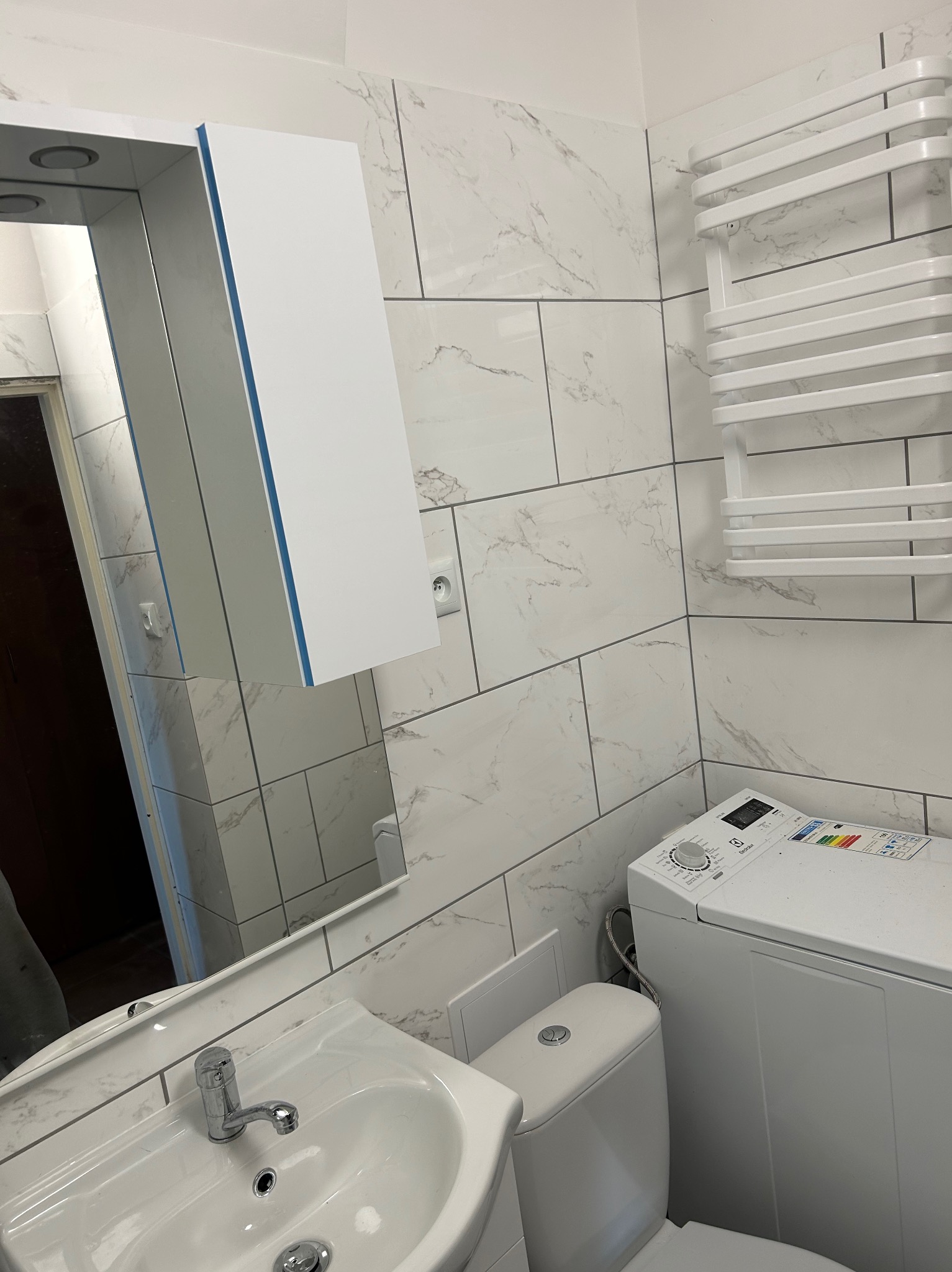 Biała łazienka z marmurowymi płytkami, umywalką, toaletą i pralką. Widoczna szafka z lustrem z niebieskim akcentem i grzejnik drabinkowy.