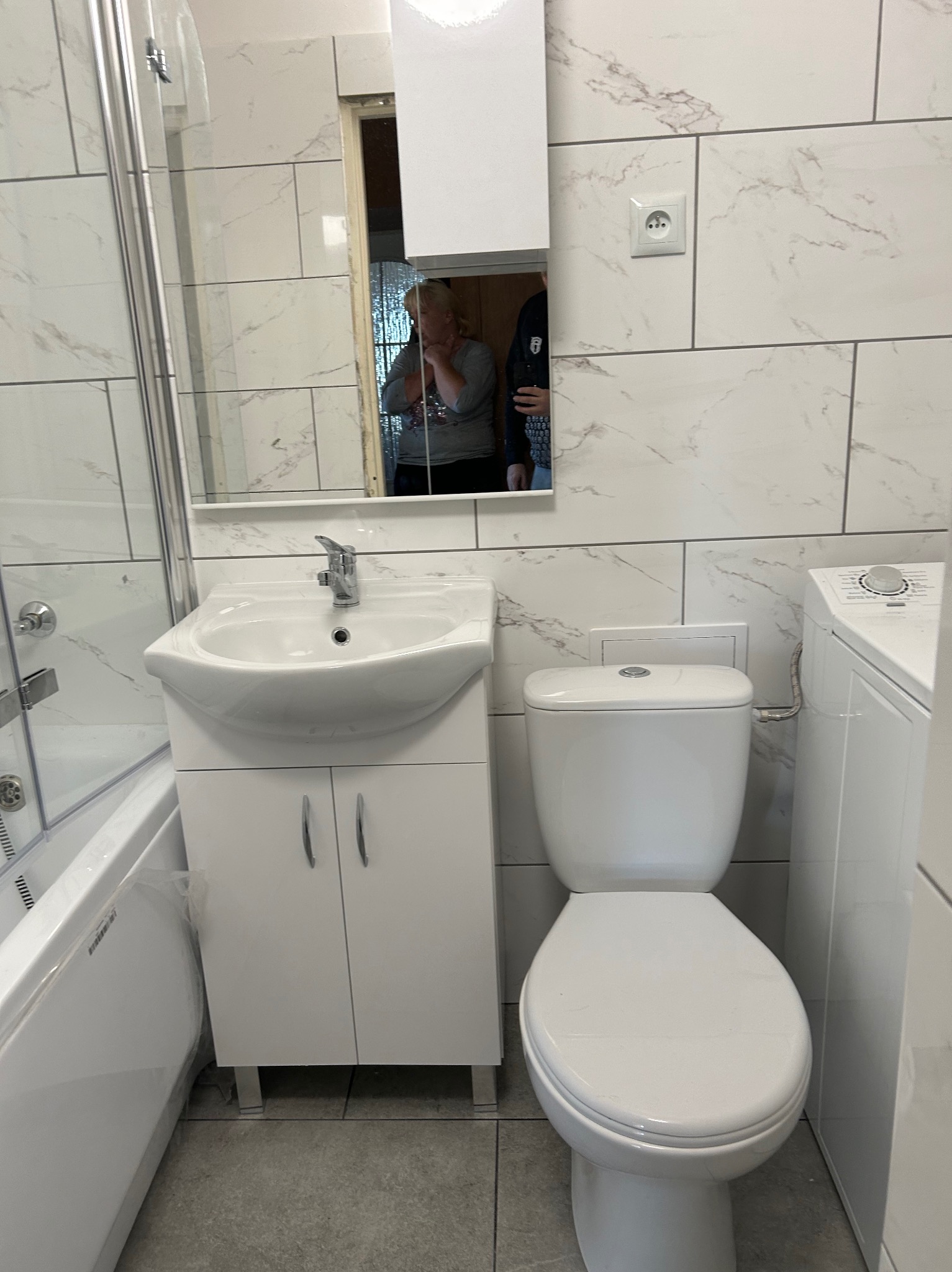 Nowoczesna, biała łazienka z marmurowymi płytkami, umywalką z szafką, toaletą i pralką, widoczna instalacja elektryczna na ścianie.