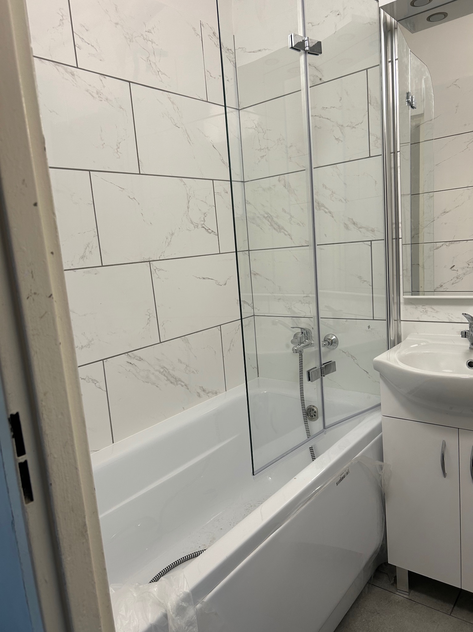 Nowa łazienka z białą wanną zabezpieczoną folią, kabiną prysznicową ze szklanymi drzwiami, umywalką z szafką i marmurkowymi płytkami na ścianach