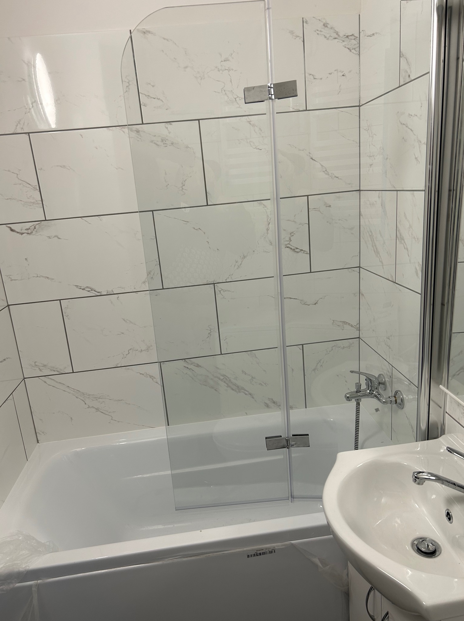 Nowoczesna łazienka z białą armaturą i szklanym parawanem nawannowym, płytki imitujące marmur na ścianach, widoczny fragment umywalki i baterii.