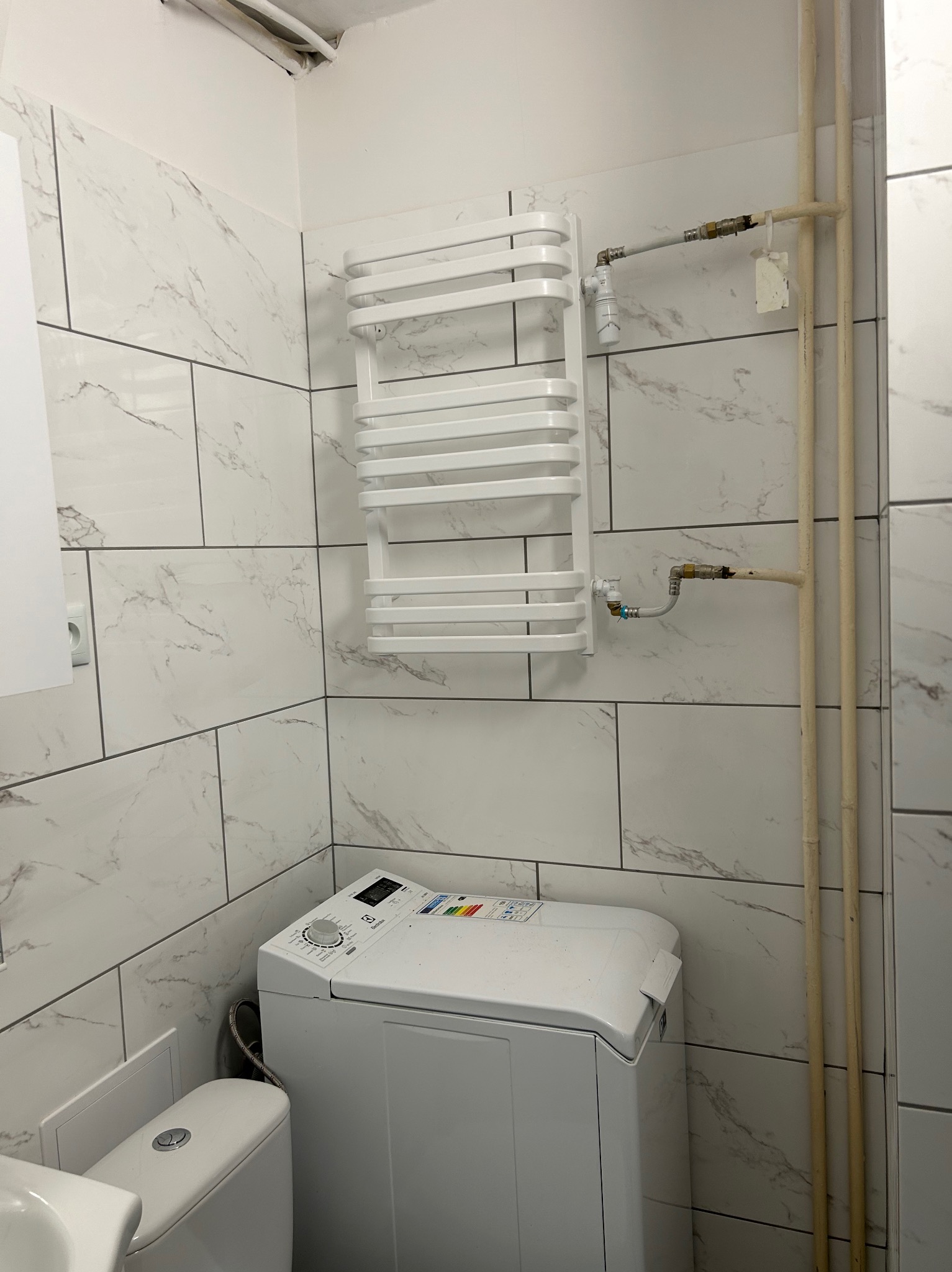 Biała łazienka z marmurkowymi płytkami, grzejnikiem drabinkowym, pralką i rurami. Widoczne przyłącza grzejnika do instalacji.