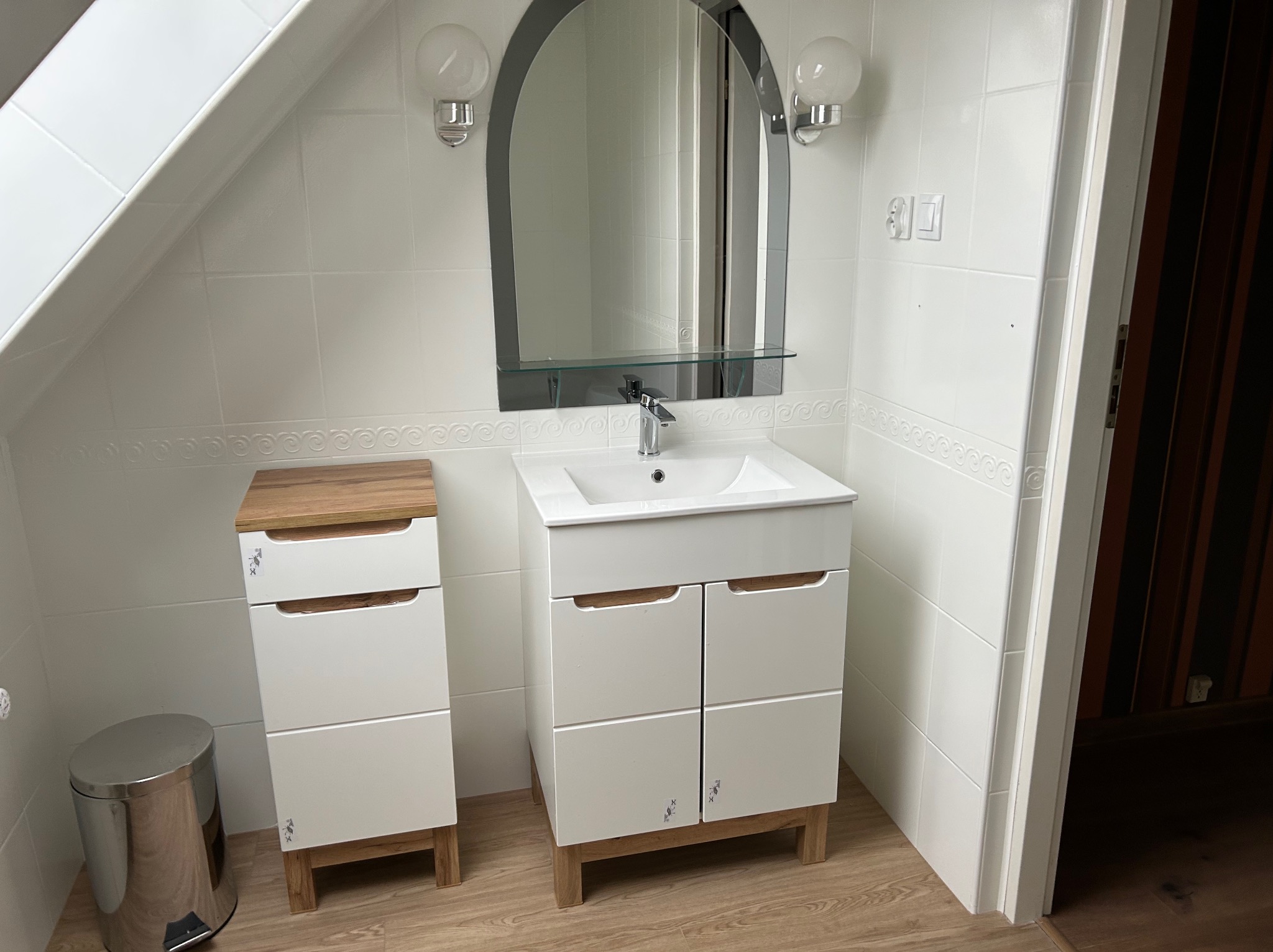 Biała łazienka na poddaszu z umywalką w szafce, lustrem w czarnej ramie i białą armaturą, wykończona białymi płytkami z dekoracyjnym paskiem i drewnopodobną podłogą.