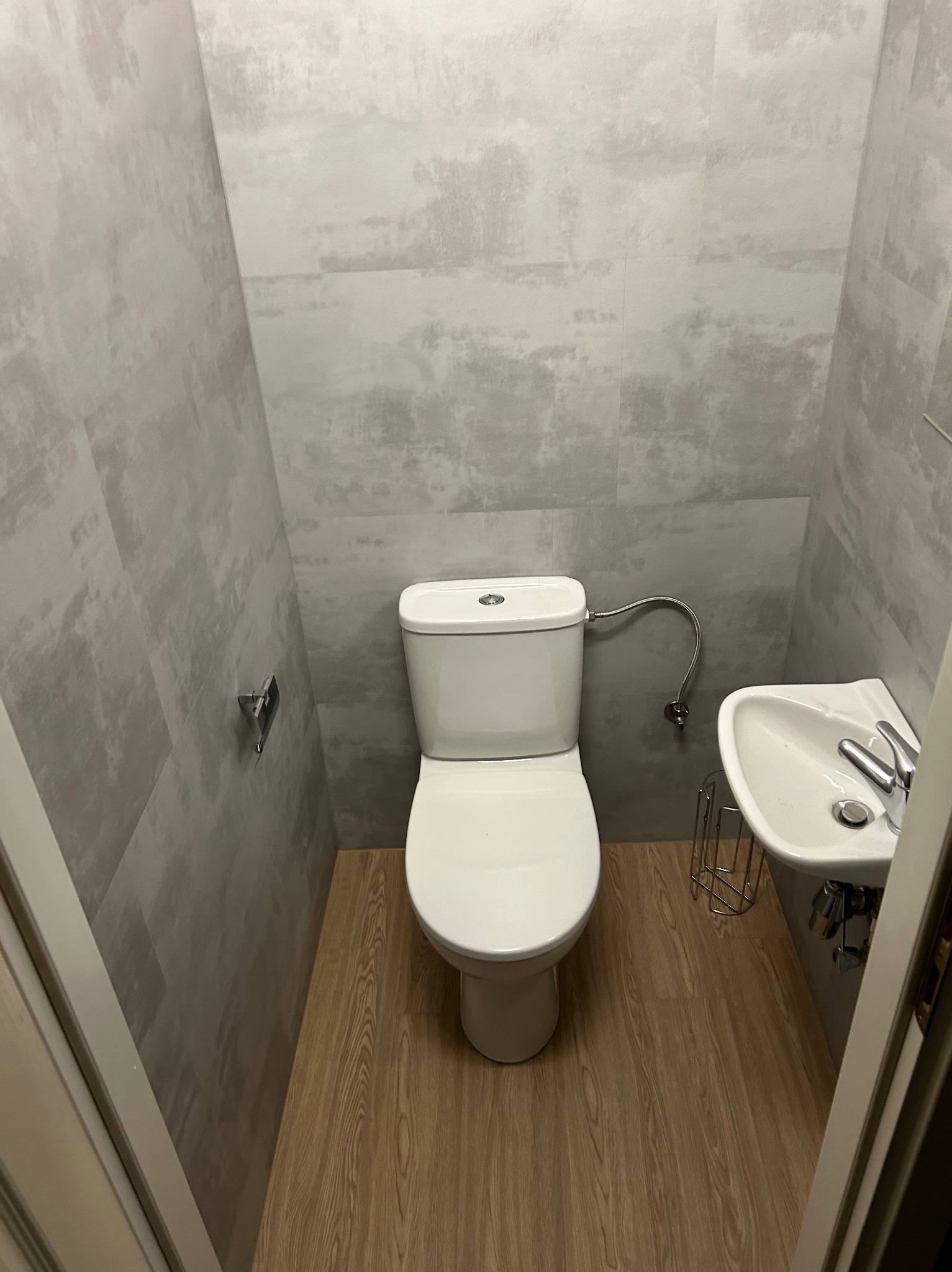 Mała toaleta z szarą, imitującą beton, okładziną ścienną i podłogą z paneli imitujących drewno. Widoczna biała muszla klozetowa i niewielka umywalka na metalowym stelażu.