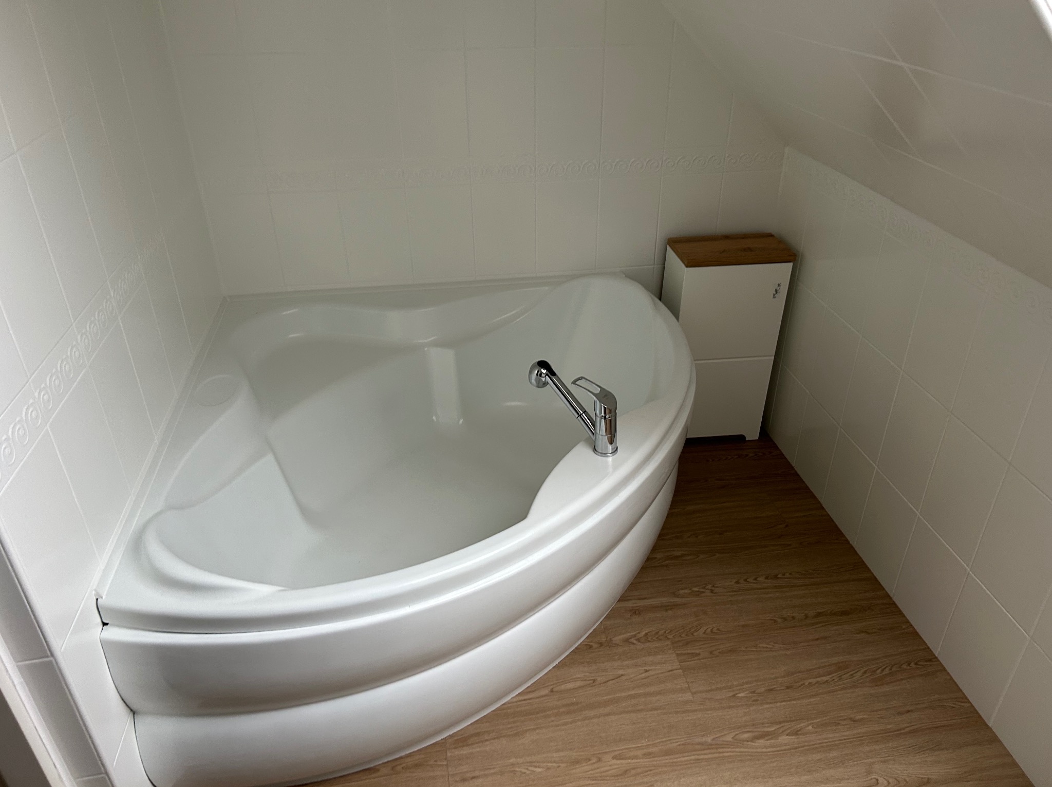 Biała narożna wanna z chromowaną baterią w łazience na poddaszu z jasnymi płytkami i drewnopodobną podłogą, obok szafka z drewnianym blatem.