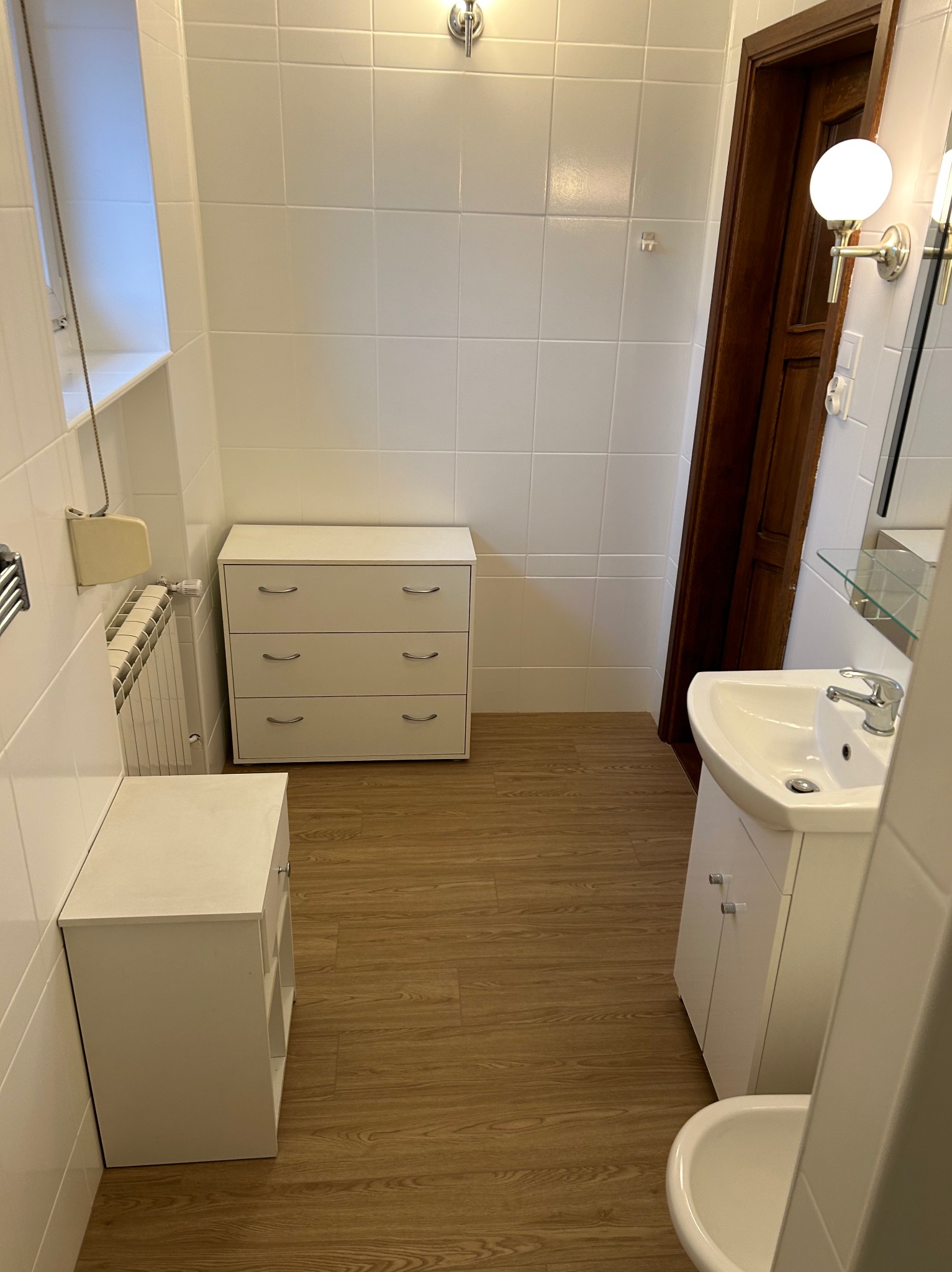 Umywalka z szafką, komoda z szufladami i grzejnik w wykończonej łazience z białymi płytkami i drewnopodobną podłogą.