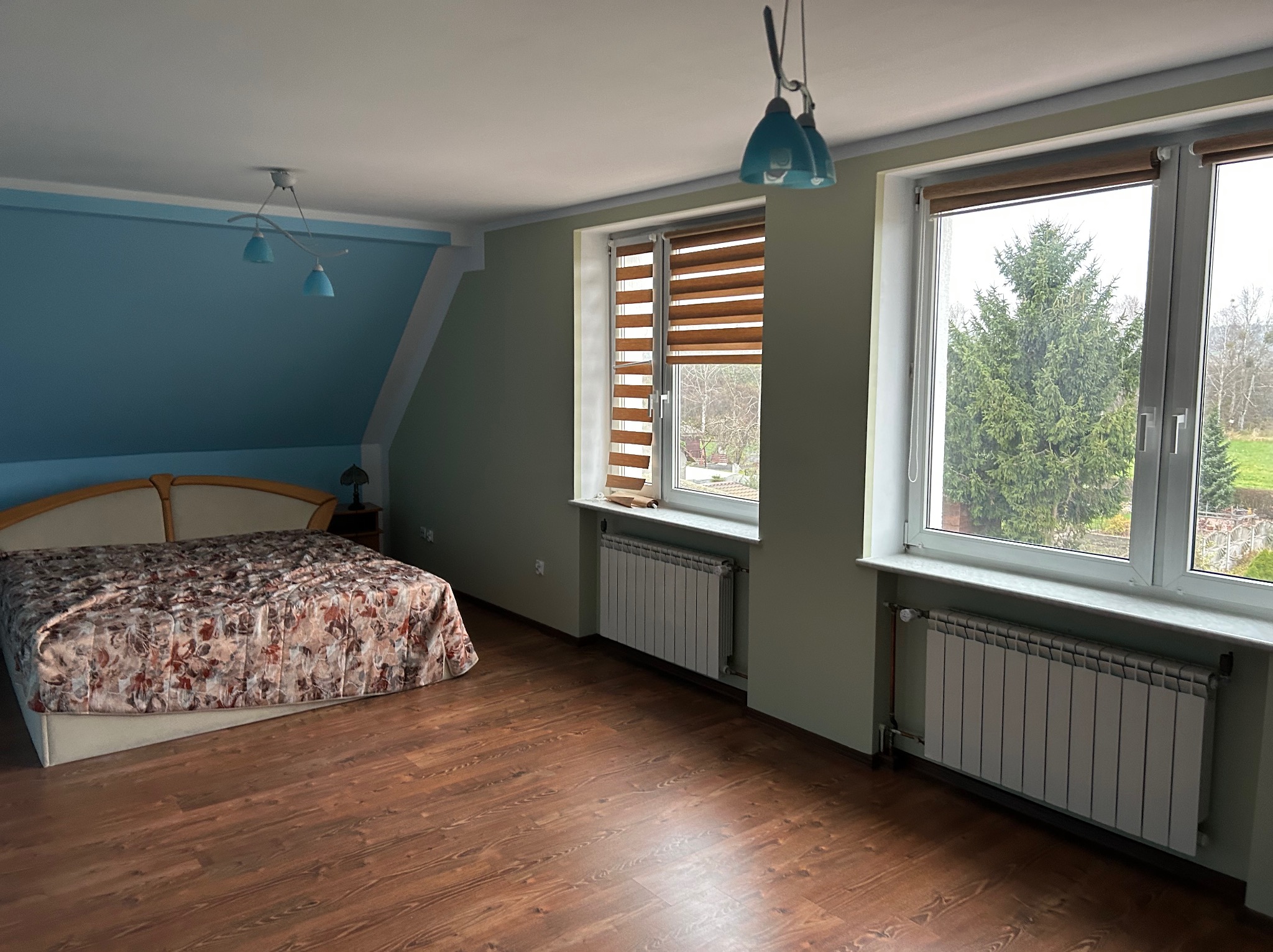 Pokój na poddaszu z drewnianą podłogą, łóżkiem z wzorzystą narzutą, oknami z widokiem na zieleń i grzejnikami pod oknami. Ściany w kolorze zielonym i niebieskim.