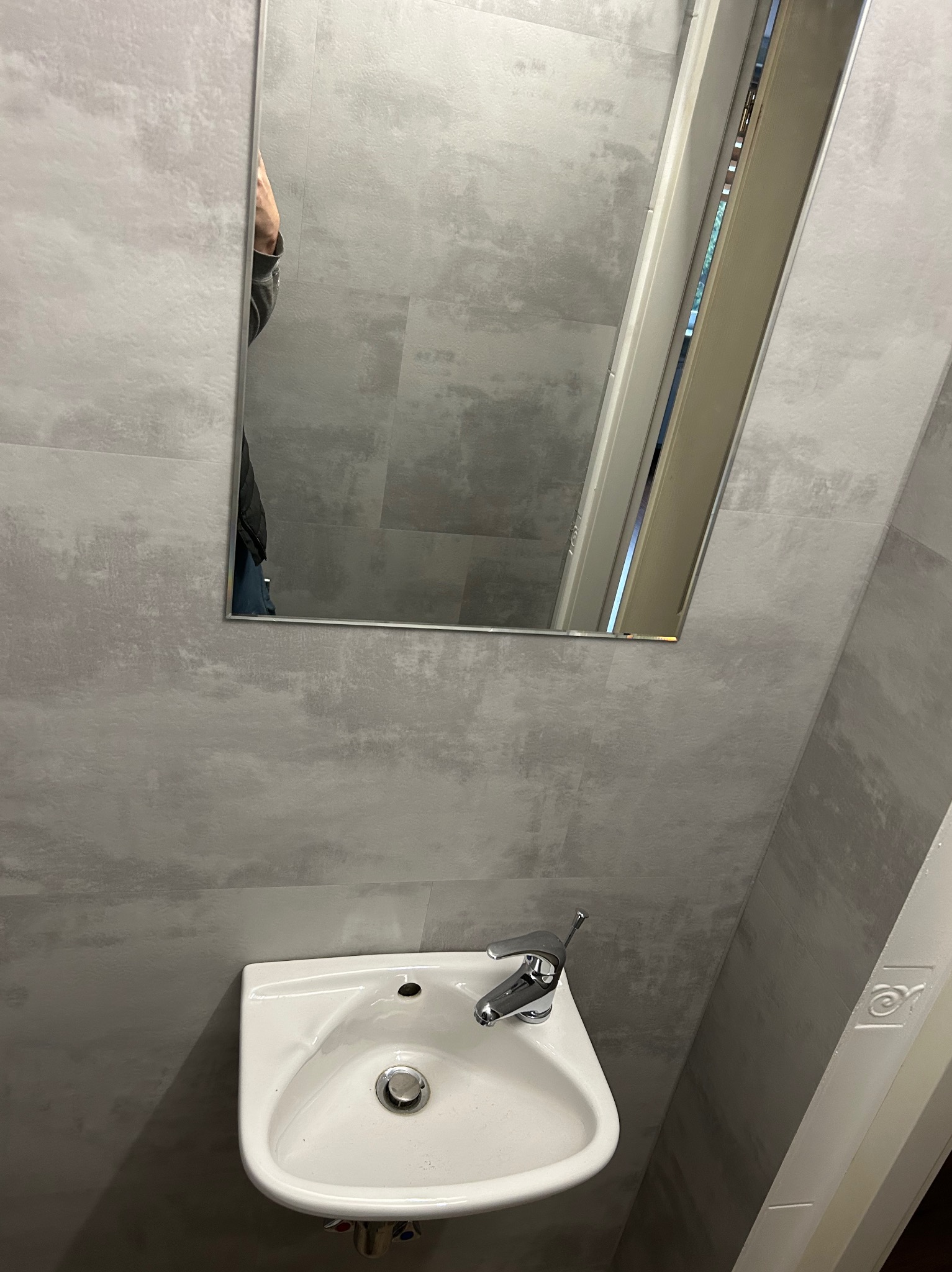 Mała łazienka z umywalką i lustrem, szare płytki imitujące beton na ścianach, minimalistyczna armatura.