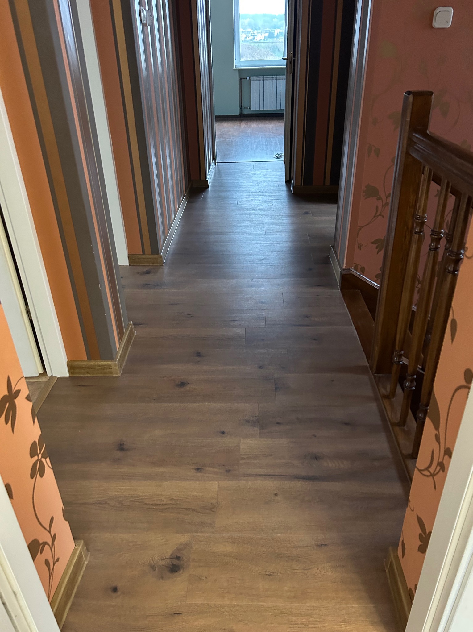 Długi korytarz z panelami podłogowymi w odcieniach brązu, ściany w paski i z kwiatowym wzorem, widok na okno na końcu korytarza.
