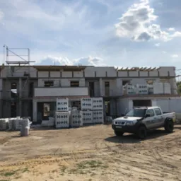 Wznoszenie dwukondygnacyjnego budynku z betonu komórkowego, widoczne palety z materiałem, rusztowanie i samochód dostawczy z przyczepką na placu budowy w słoneczny dzień.