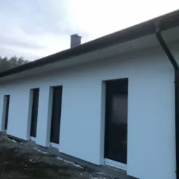 Świeżo otynkowany, parterowy budynek jednorodzinny z ciemnymi oknami i orynnowaniem, widoczny komin i fragment terenu przed budynkiem.