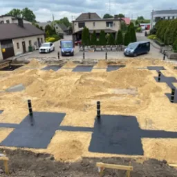 Przygotowanie terenu pod budowę: wykopane fundamenty, czarne folie izolacyjne, drewniane szalunki, widoczne słupki pionowe, w tle budynki mieszkalne i zaparkowane samochody.