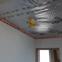 Budmar Marcin Garncarek - Izolacja termiczna poddasza z folią aluminiową i wełną mineralną, widoczny kabel elektryczny i fragment okna w surowym stanie.