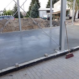 Budmar Marcin Garncarek - Świeżo wylana, gładka betonowa posadzka hali z widocznymi metalowymi słupami konstrukcyjnymi i stertą ziemi w tle, obok stoi miotła.