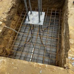 Zbrojenie fundamentu z wystającymi prętami i stalową płytą kotwiącą, umieszczone w wykopie.