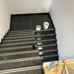 Budmar Marcin Garncarek - Schody wyłożone ciemnym gresem z białymi paskami antypoślizgowymi, widoczne narzędzia i materiały budowlane, etap prac wykończeniowych.