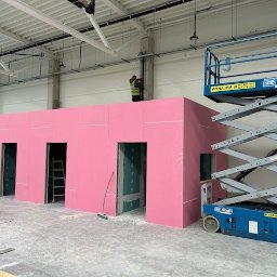 Budmar Marcin Garncarek - Wnętrze hali z budową ścian działowych z różowych płyt gipsowo-kartonowych, widoczny podnośnik nożycowy i pracownicy na rusztowaniu.